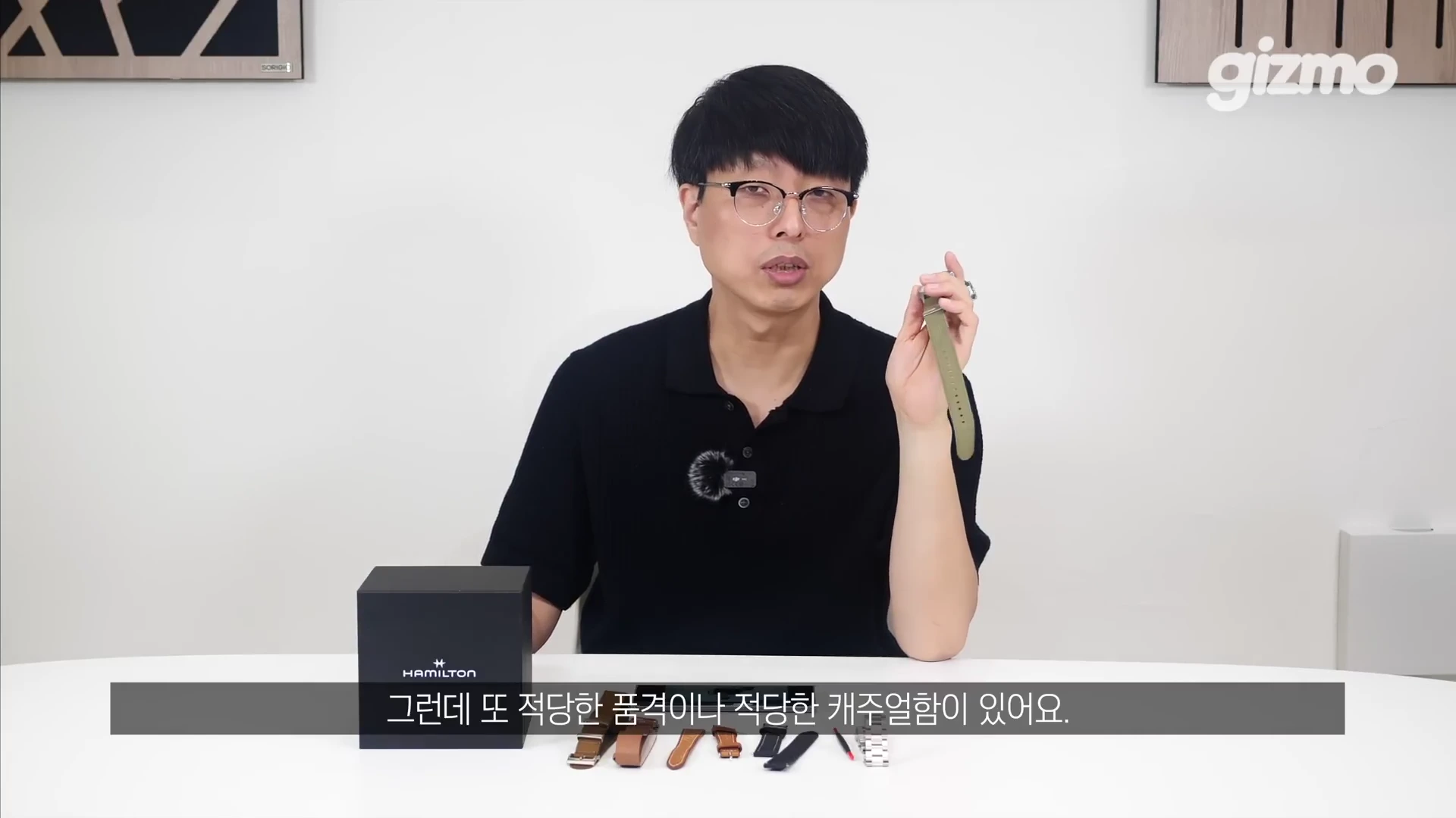 흰색 배경의 스튜디오에서 안경을 쓴 남성이 한 손에 올리브색 시계 스트랩을 들어 보이고 있으며, 테이블 위에는 검은색 해밀턴 시계 상자와 다양한 교체용 시계줄들이 놓여 있습니다.