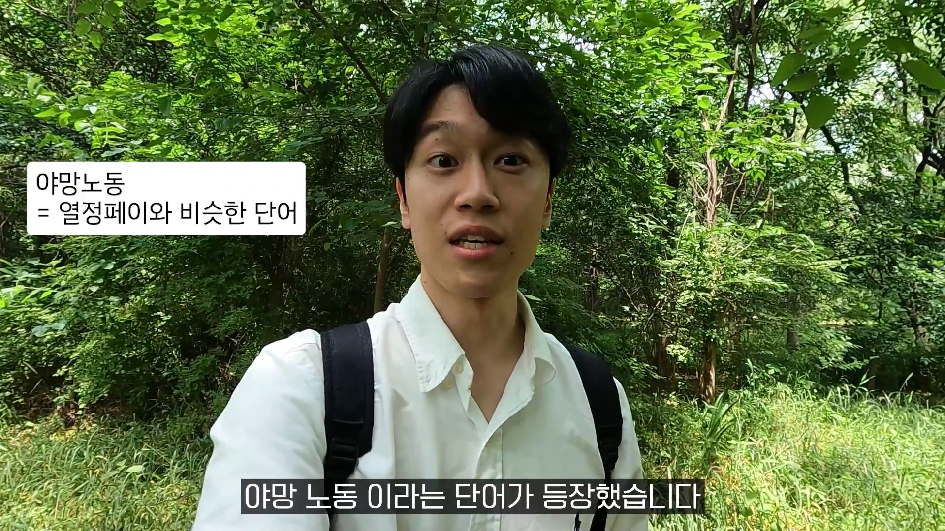 흰색 셔츠를 입고 백팩을 멘 남성이 숲속에서 카메라를 정면으로 응시하며 말하고 있으며, 화면 왼쪽 상단에는 '야망노동 = 열정페이와 비슷한 단어'라는 자막이, 하단에는 '야망 노동이라는 단어가 등장했습니다'라는 자막이 표시되어 있습니다.