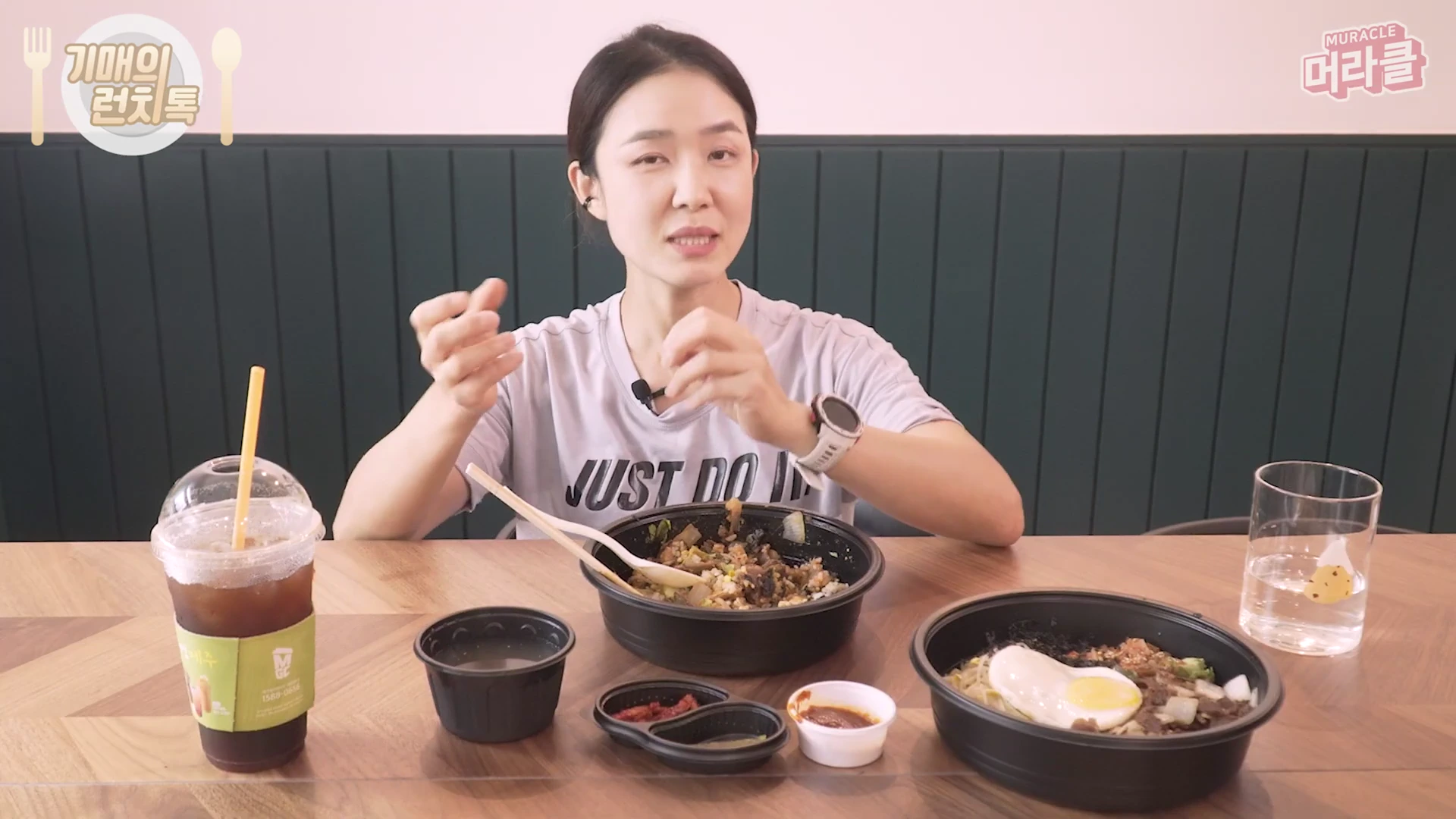 테이블 앞에 앉아 있는 여성이 손동작을 하며 무언가를 설명하고 있고, 앞에는 비빔밥과 아이스 아메리카노 등 식사 메뉴가 놓여 있다.