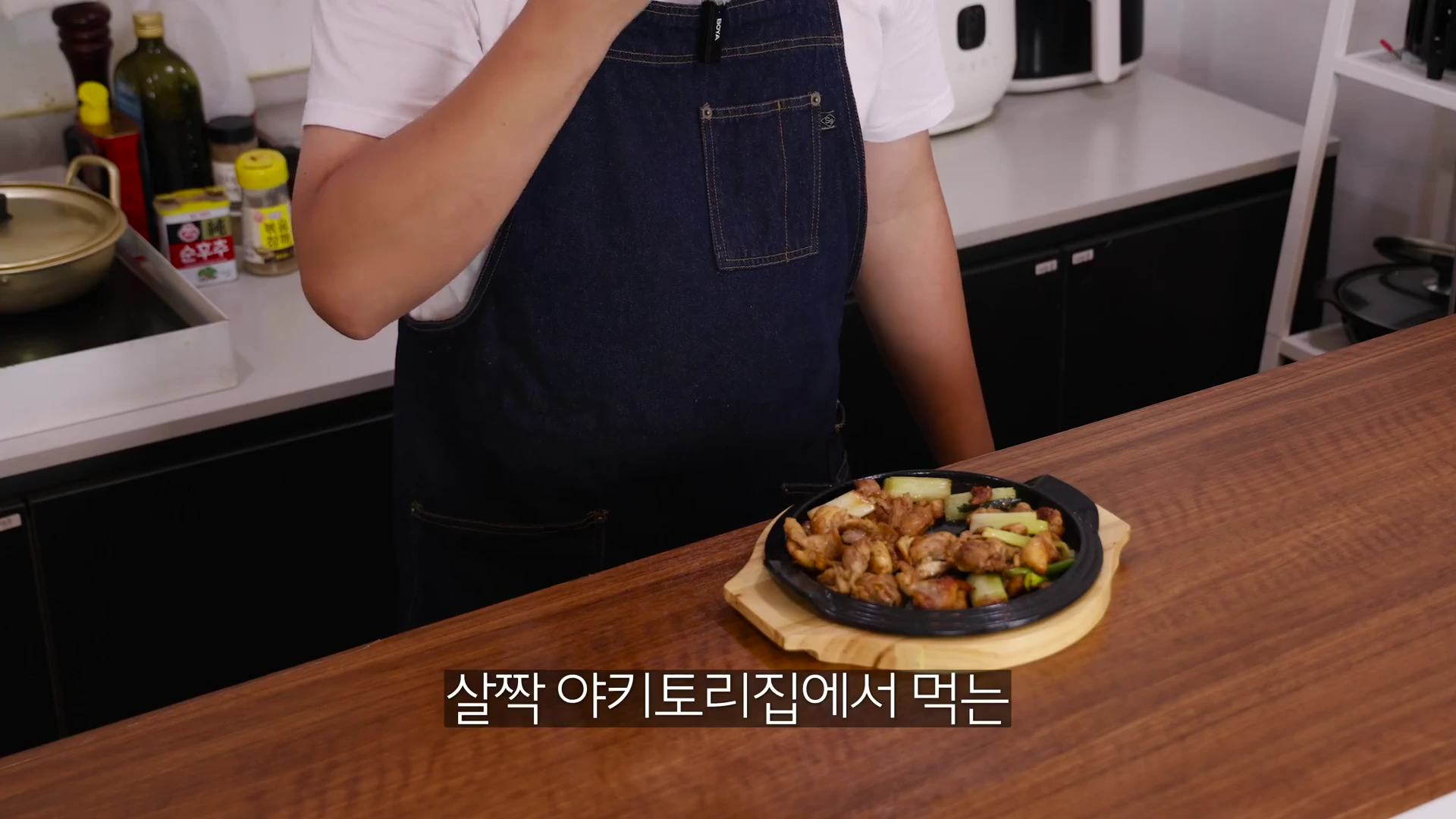 앞치마를 두른 요리사가 주방 조리대 위에 올려진 철판 위 대파 닭고기 구이를 바라보며 시식하는 모습.