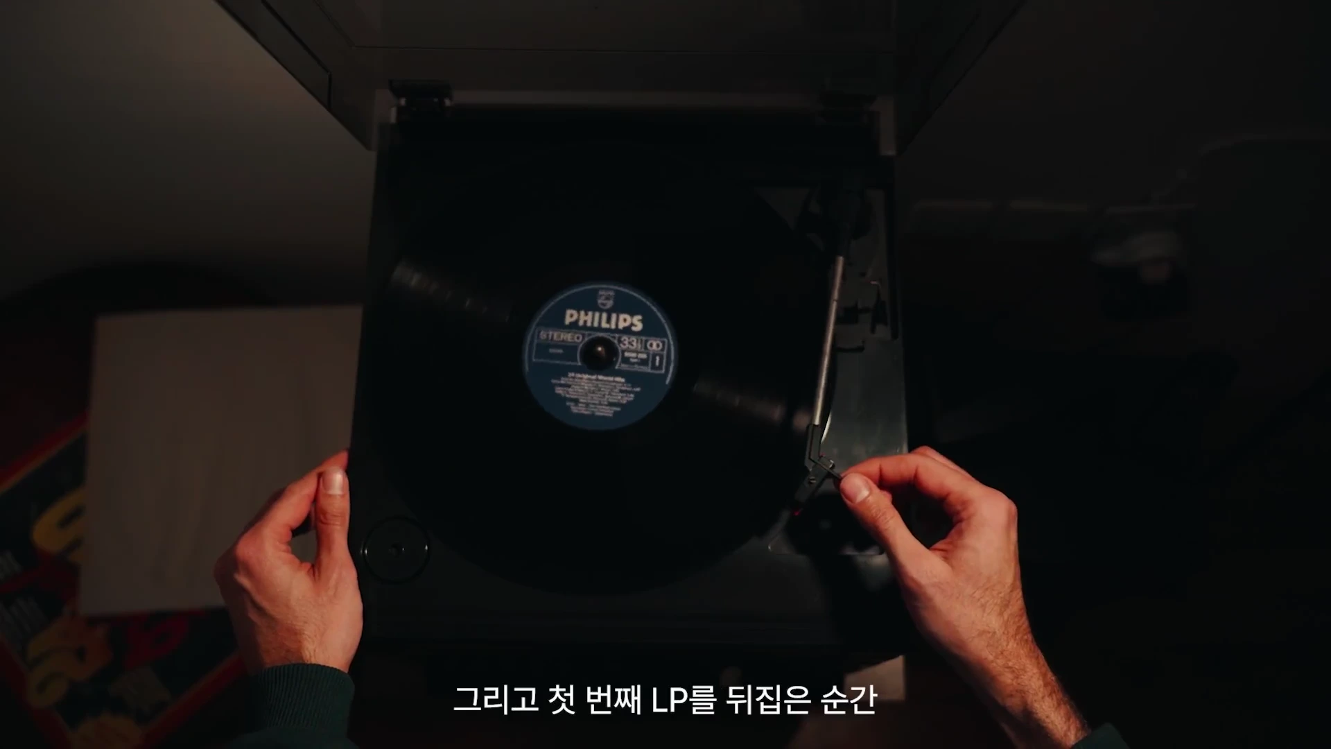 두 손으로 LP 플레이어의 톤암을 조절하여 검은색 레코드판 위에 올리는 모습이 위에서 내려다본 시점으로 촬영되었습니다.