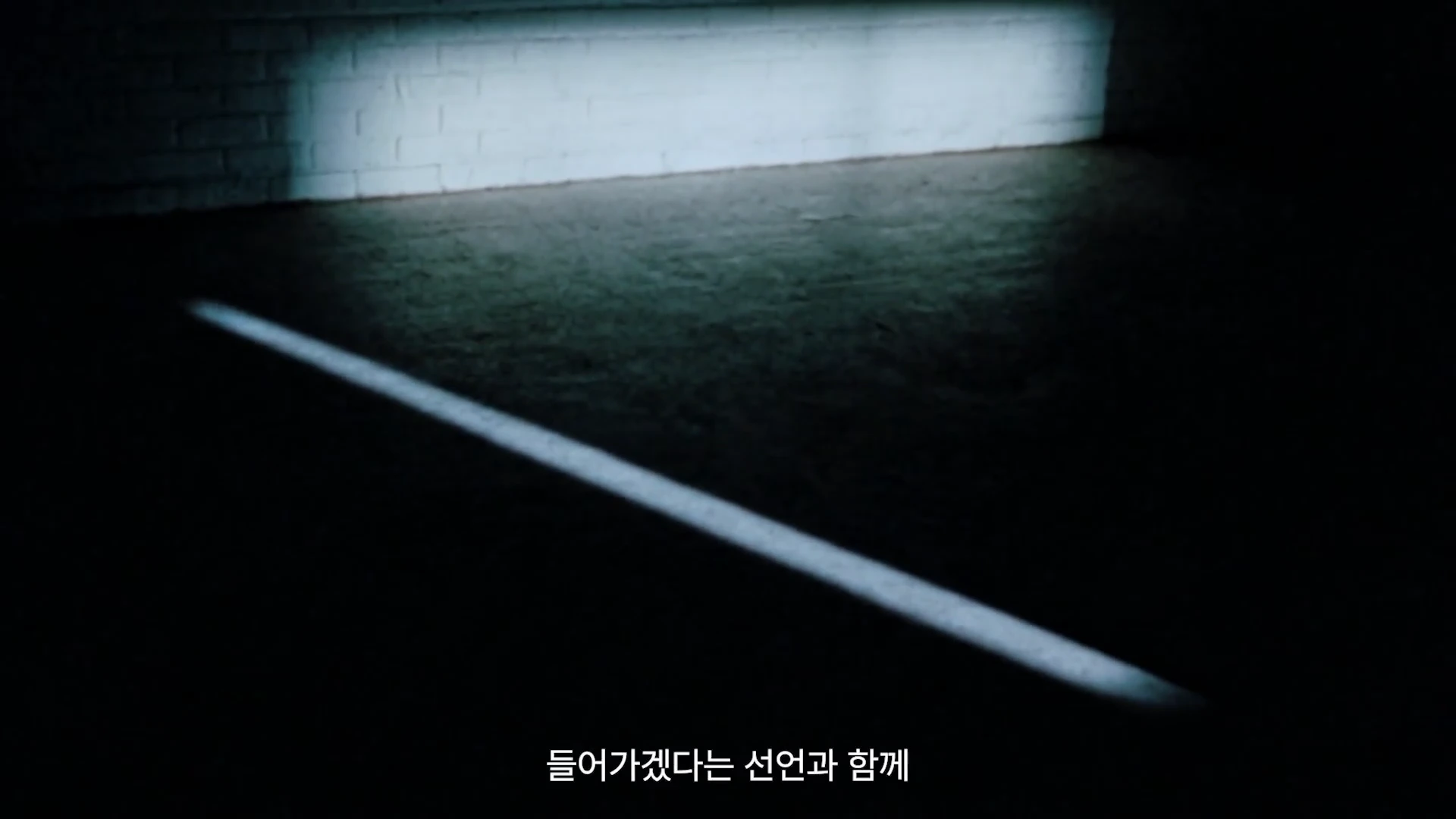 어두운 공간에 희미한 조명이 흰색 벽돌 벽의 일부를 비추고 있으며, 바닥에는 긴 직선 형태의 빛이 드리워져 있다.