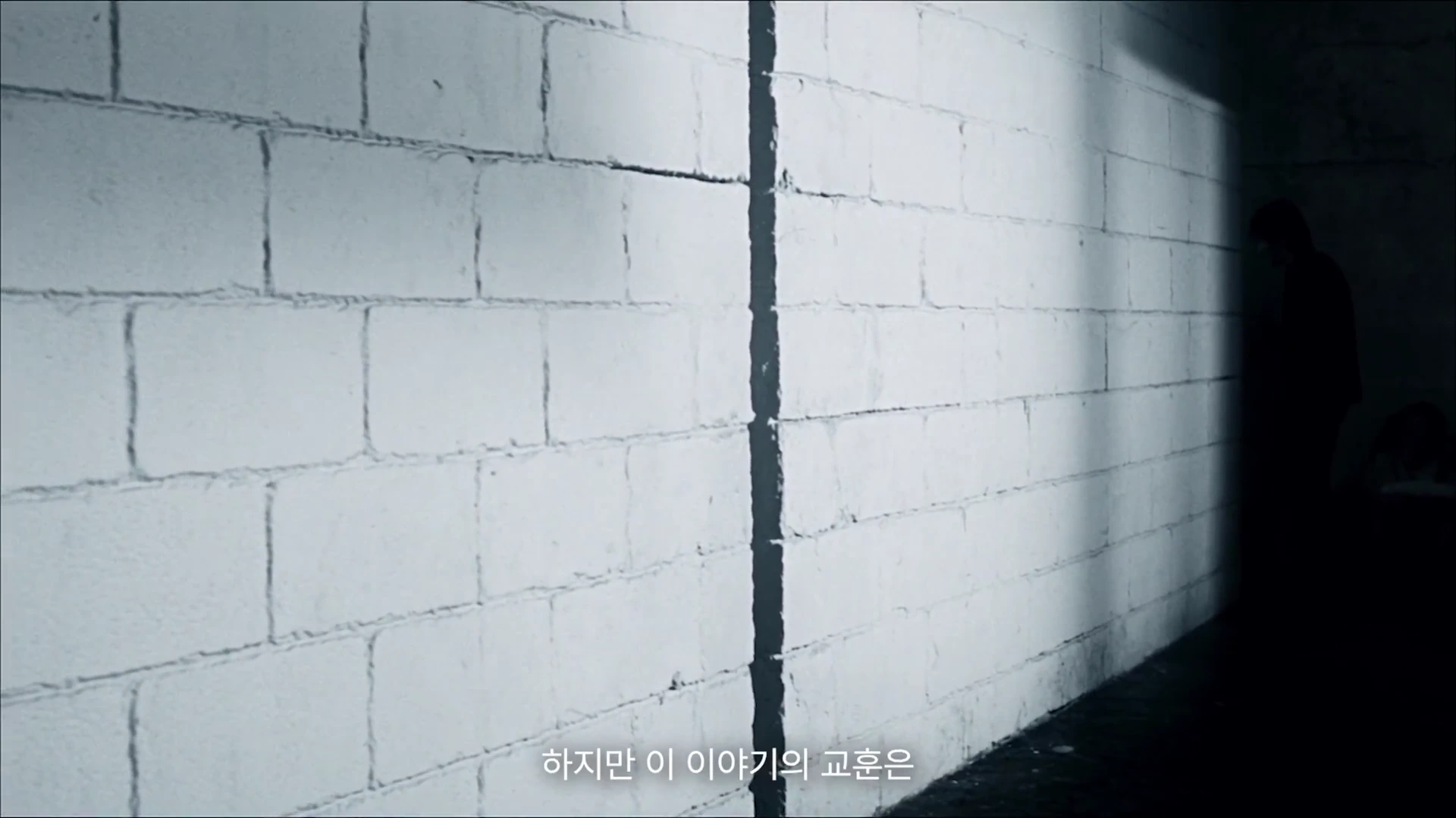 흰색 콘크리트 벽 앞에 한 남자가 어두운 실루엣으로 서 있는 영상 장면