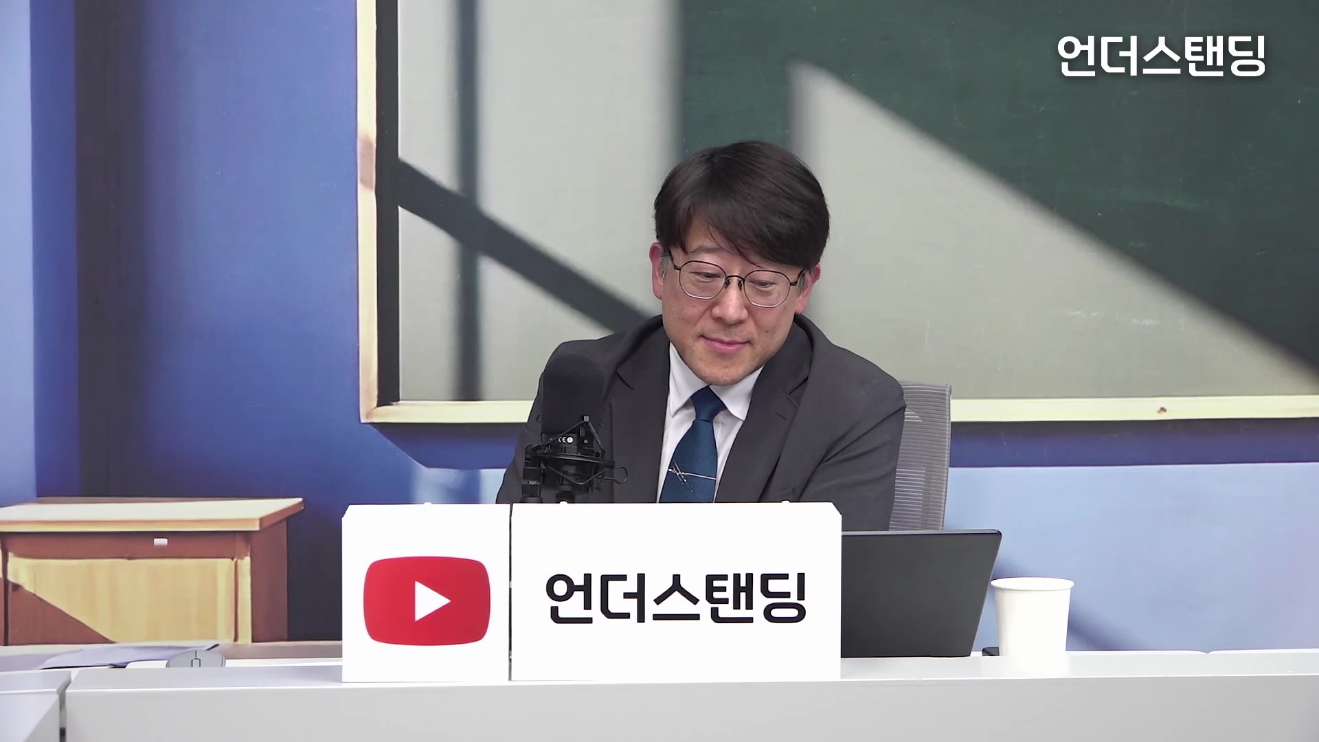 안경을 쓴 중년 남성이 정장을 입고 스튜디오 테이블에 앉아 인터뷰를 진행하고 있는 모습입니다. 앞에는 마이크와 '언더스탠딩' 로고가 적힌 표지판이 놓여 있습니다.