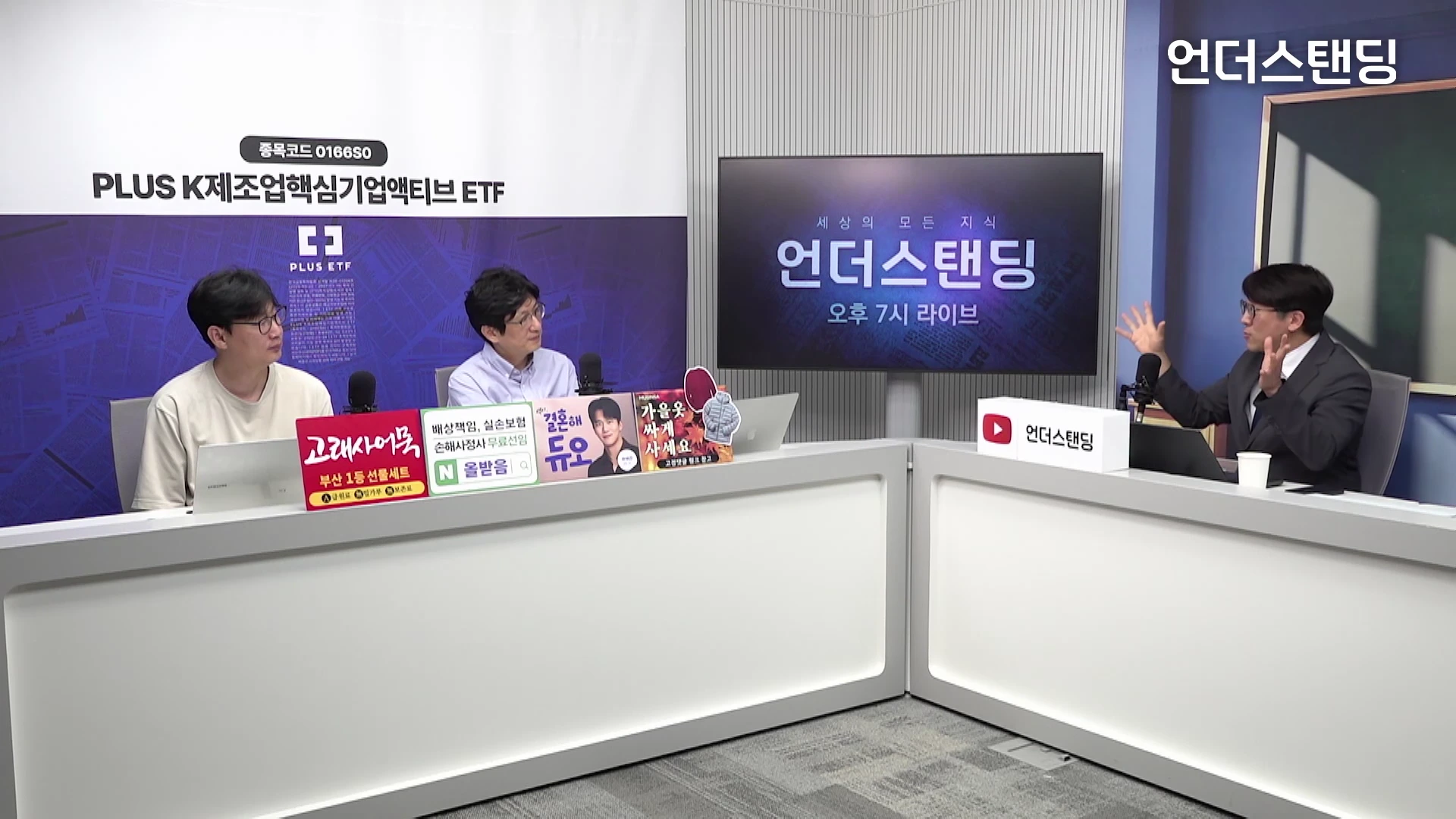 세 명의 남자가 스튜디오에서 테이블을 사이에 두고 앉아 대화를 나누고 있는 모습으로, 중앙의 화면에는 프로그램 로고가 떠 있고 테이블 위에는 다양한 안내 문구가 부착되어 있습니다.