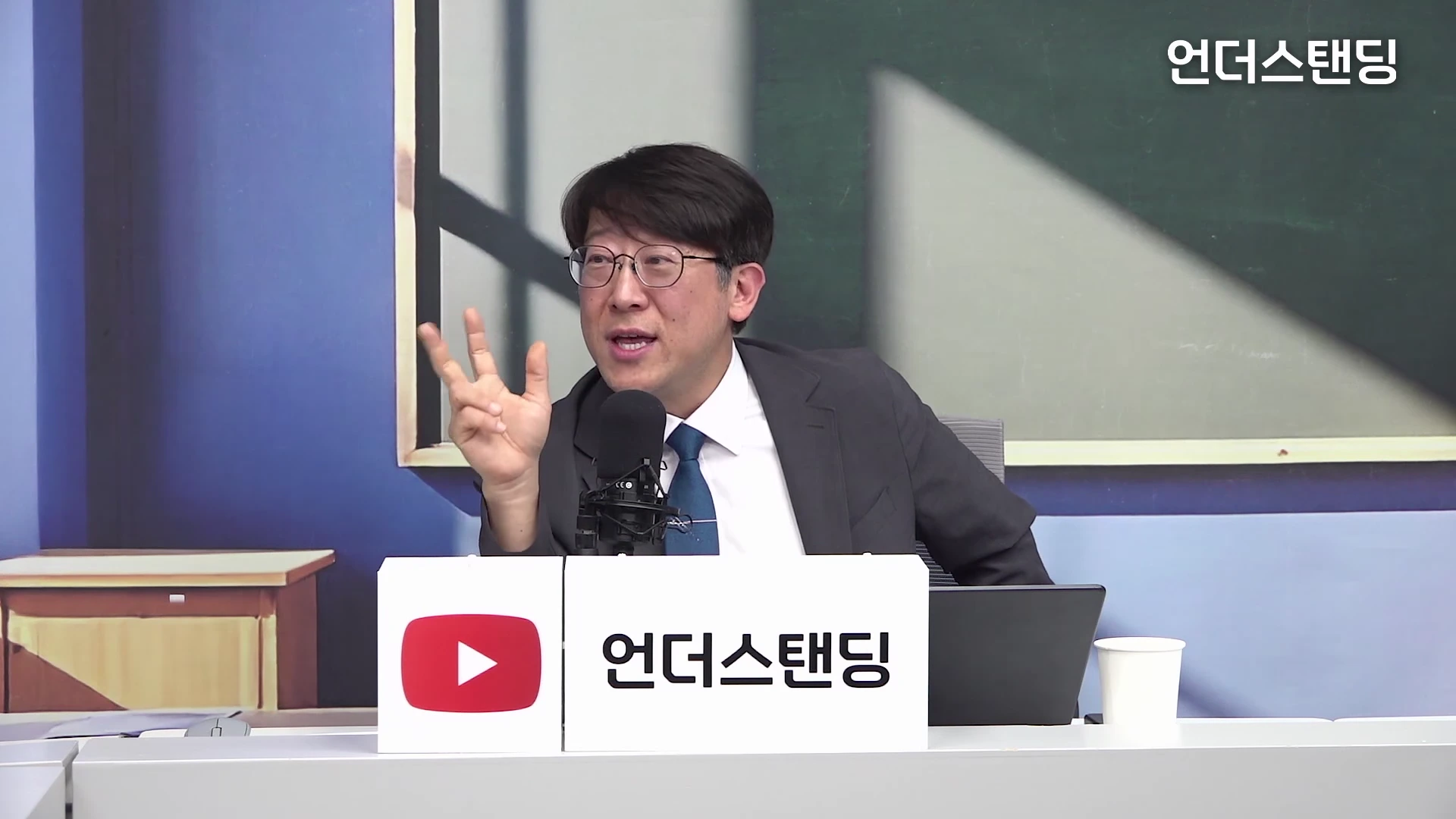 안경을 쓴 남성이 스튜디오에 앉아 손가락 세 개를 펴서 숫자를 설명하며 이야기하고 있는 모습