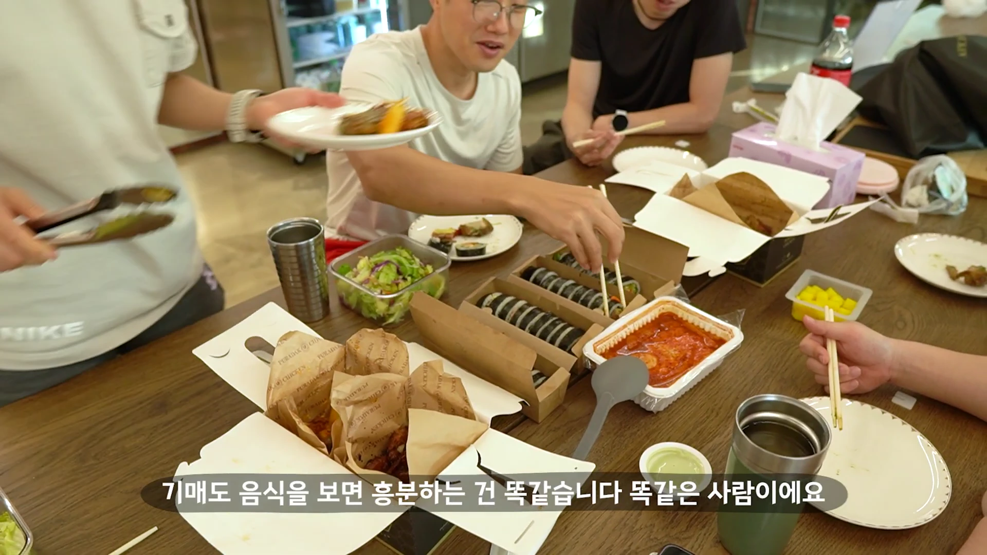 사람들이 여러 명이 모여 앉은 테이블 위에 김밥, 떡볶이, 치킨 등 다양한 배달 음식이 차려져 있고 젓가락으로 음식을 집는 모습이 담긴 화면 하단에 자막이 적힌 영상 캡처 이미지입니다.
