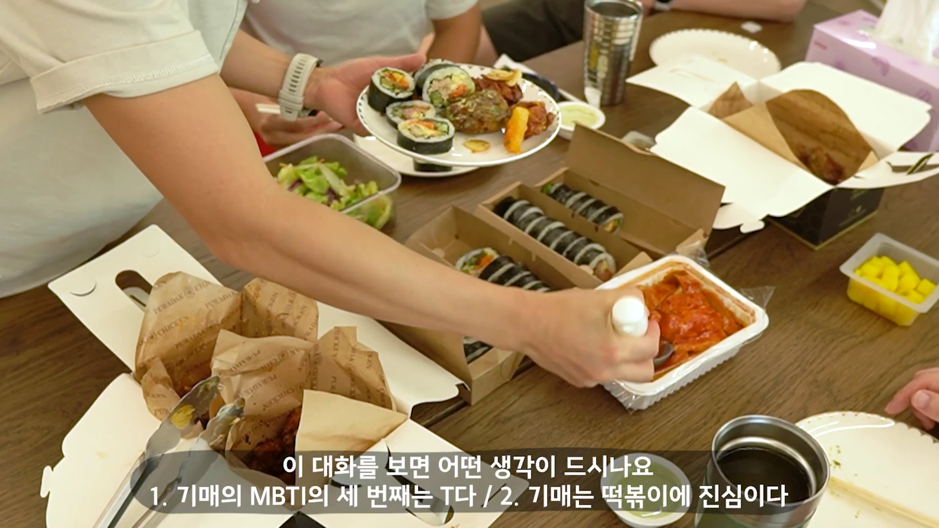 사람들이 식탁 위에 김밥, 떡볶이, 치킨, 샐러드 등 다양한 배달 음식을 차려놓고 식사 준비를 하는 모습