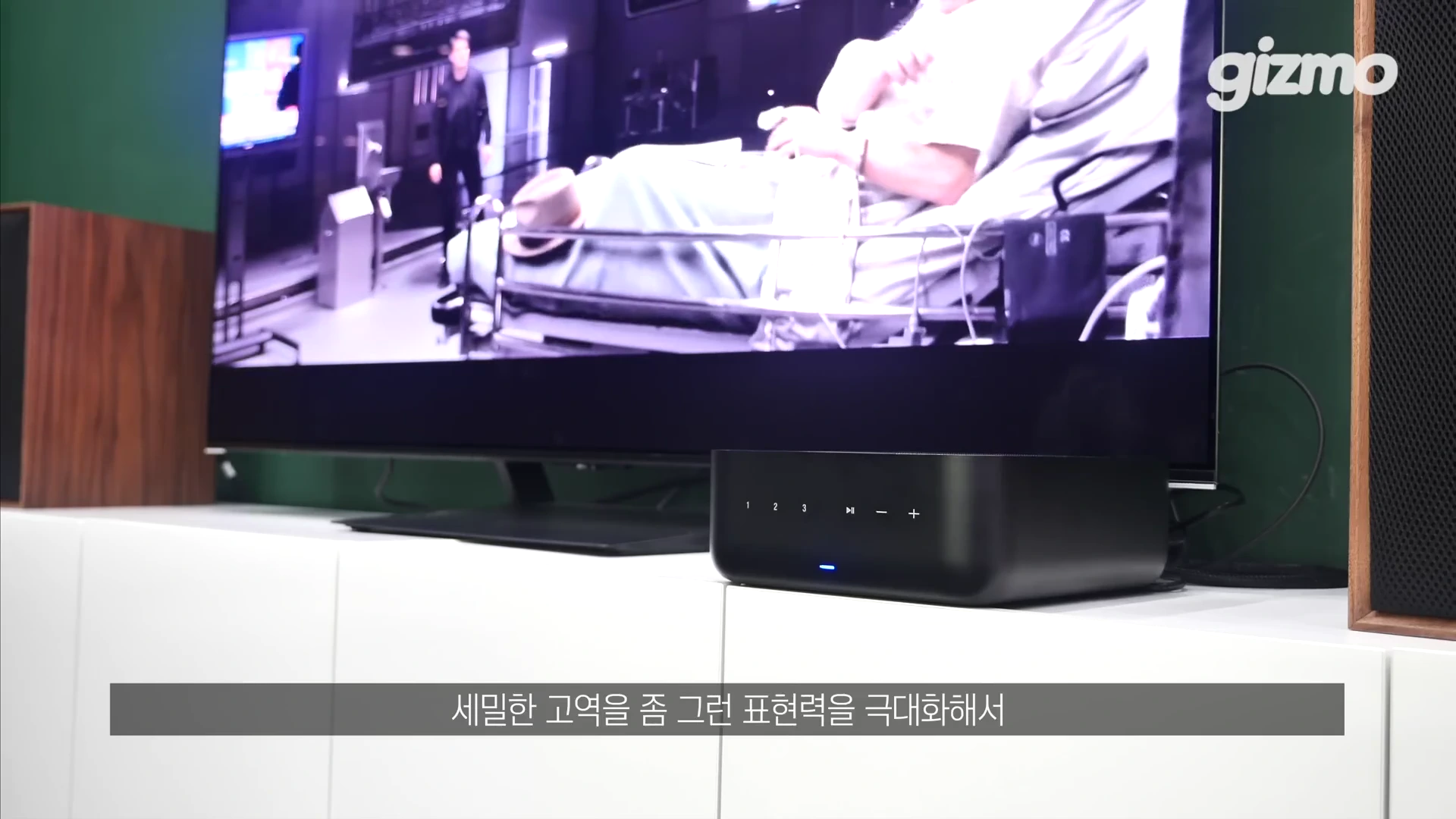 흰색 가구 위에 놓인 검은색 앰프와 그 뒤로 TV 화면이 보이는 거실 공간.