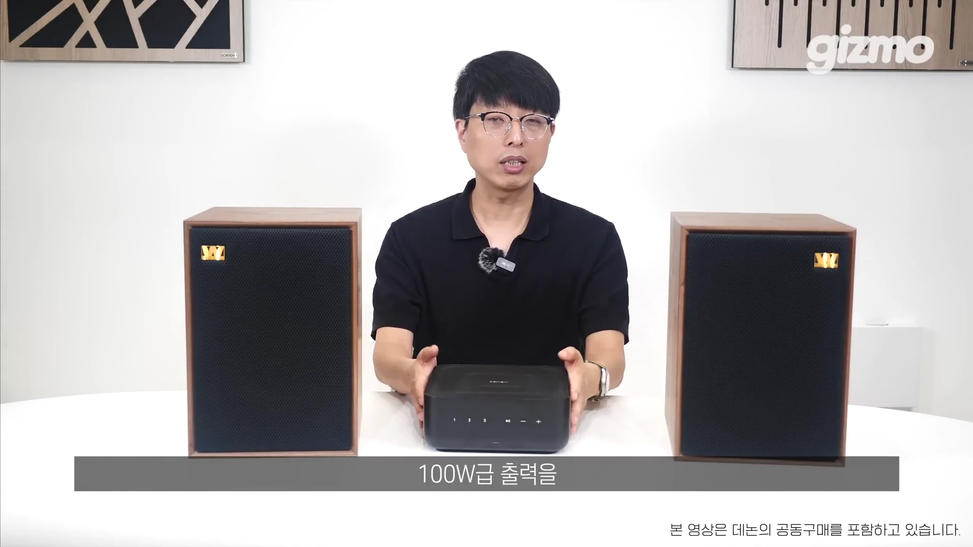 흰색 배경의 스튜디오에서 안경을 쓴 남성이 양쪽에 스피커를 두고 중앙에서 앰프를 두 손으로 잡고 설명하는 모습
