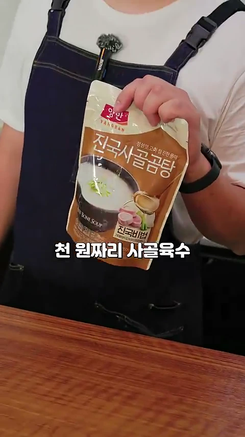 앞치마를 입은 사람이 한 손으로 시판용 파우치 형태의 사골곰탕 제품을 들어 보이고 있다.