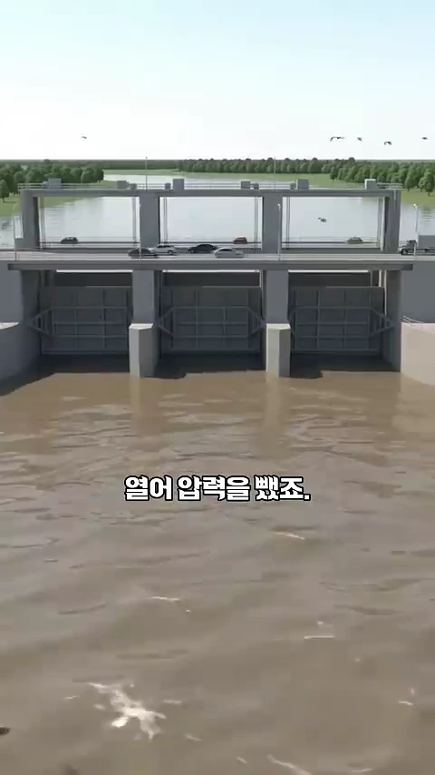 회색 콘크리트로 지어진 대형 수문이 강을 가로지르고 있으며, 수문 위로는 차량이 다니는 도로가 있고 중앙에는 '열어 압력을 뺐죠'라는 자막이 적힌 3D 그래픽 영상 화면입니다.