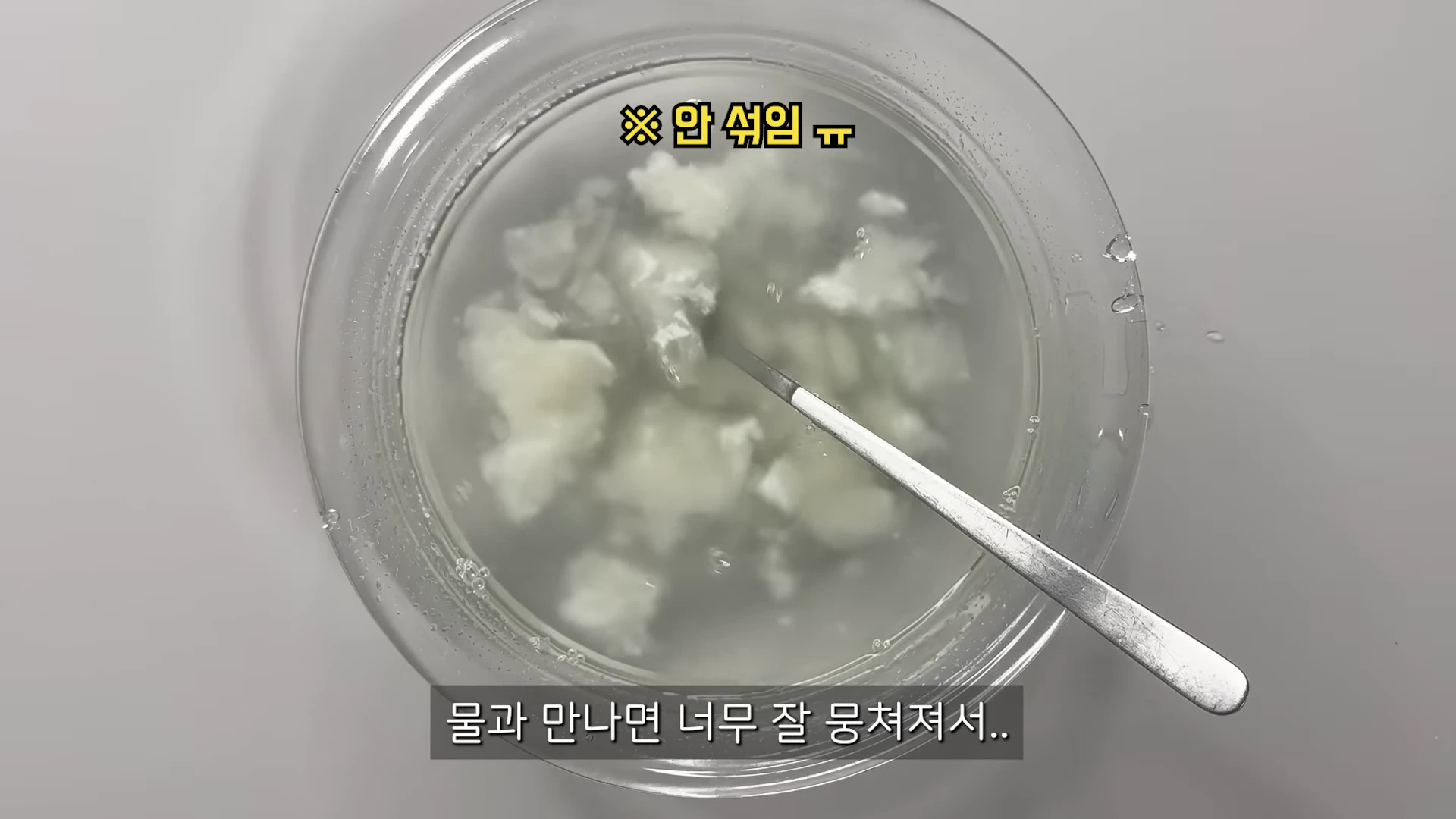 투명한 유리 볼 안에 물과 섞이지 않고 덩어리진 하얀 가루가 담겨 있으며, 그 위에는 숟가락이 놓여 있습니다.
