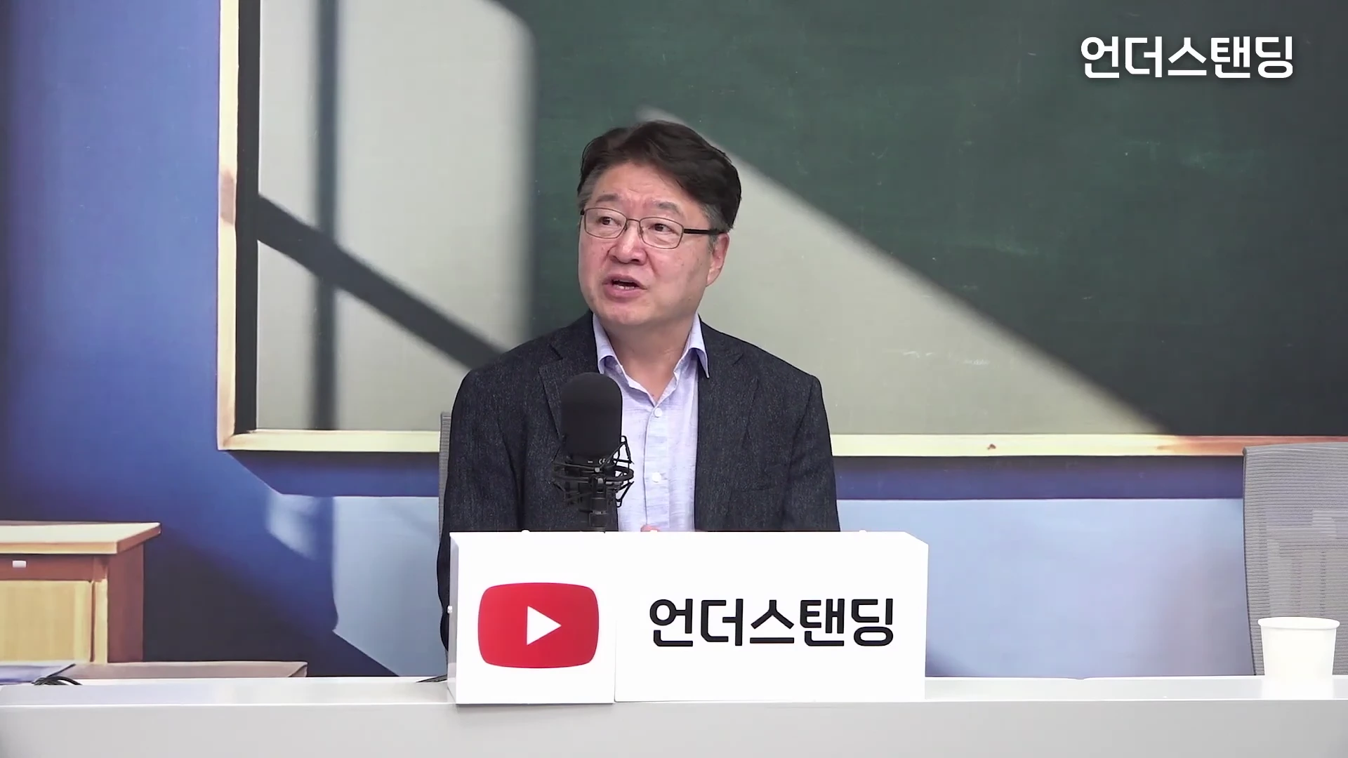 안경을 쓴 중년 남성이 테이블에 놓인 마이크 앞에서 진지한 표정으로 이야기를 하고 있는 모습이다.