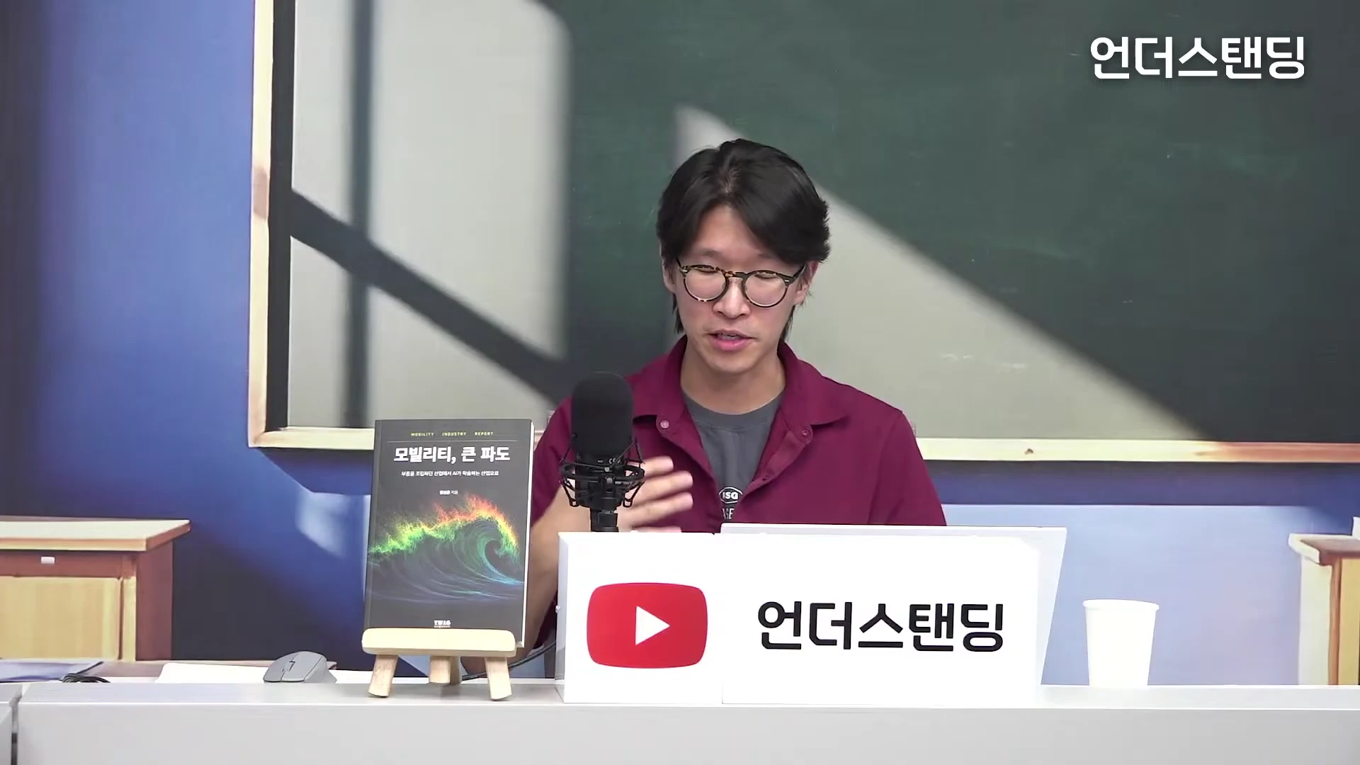 안경을 쓴 한 남성이 스튜디오 책상에 앉아 손짓하며 이야기를 나누고 있다. 옆에는 '모빌리티, 큰 파도'라는 제목의 책이 거치대에 놓여 있다.