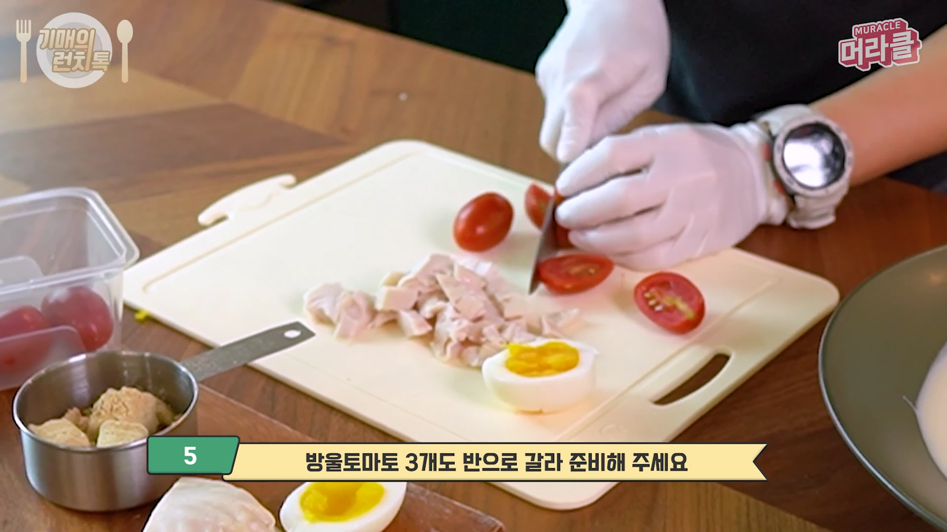 흰색 위생 장갑을 낀 손이 도마 위에서 방울토마토를 칼로 반으로 자르고 있고, 옆에는 삶은 달걀과 잘게 썬 닭가슴살이 놓여 있습니다.