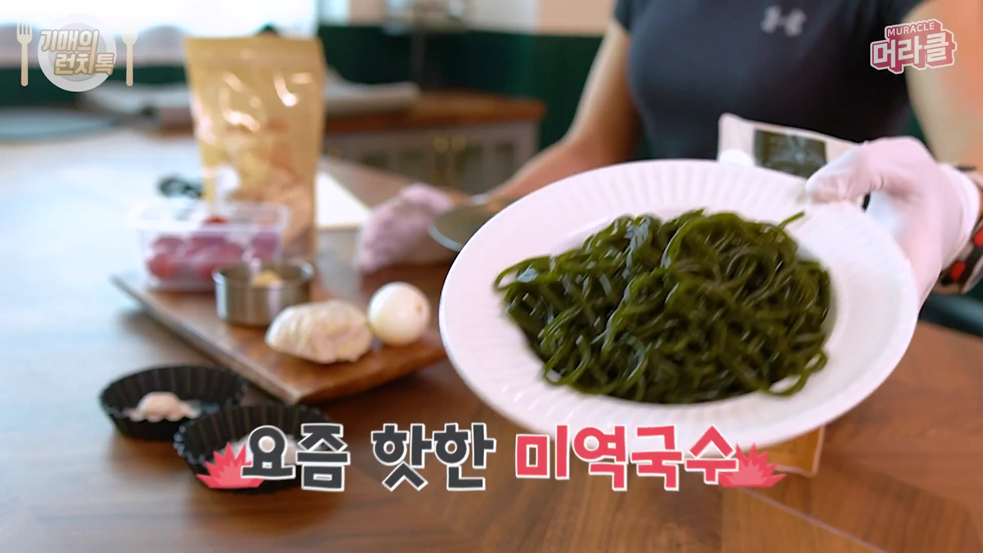 흰 접시에 담긴 초록색 미역국수를 한 손으로 들고 있는 모습과 그 뒤로 콩물과 닭가슴살 등 재료가 놓여 있는 테이블이 보입니다.