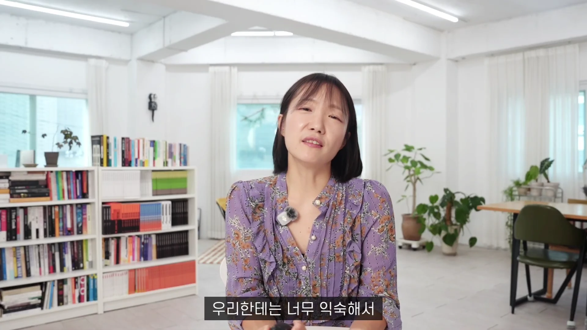 단발머리를 한 여성이 밝은 실내에서 정면을 바라보며 이야기하고 있고, 뒤쪽으로는 책이 가득 꽂힌 책장과 화분이 놓여 있다.