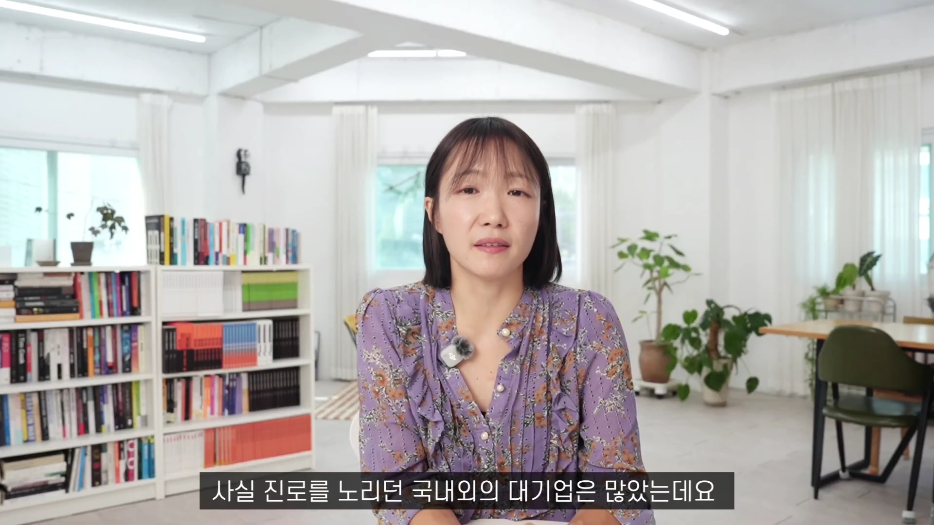 단발머리를 한 여성이 꽃무늬 블라우스를 입고 밝은 실내 공간에서 정면을 응시하며 이야기하고 있다. 배경에는 책장이 놓여 있고 하단에는 '사실 진로를 노리던 국내외의 대기업은 많았는데요'라는 자막이 표시되어 있다.