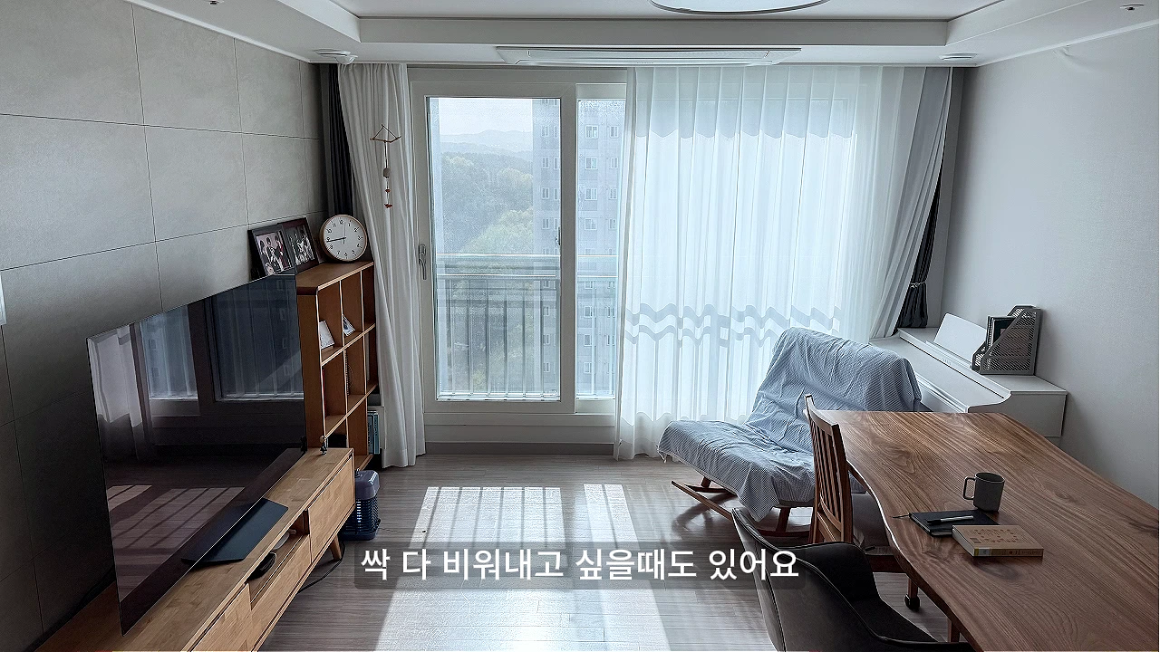 창가에 밝은 빛이 들어오는 차분한 분위기의 거실에 나무 소재의 거실장과 식탁, 안락의자가 배치되어 있고 하단에 자막이 있는 영상 캡처 화면.