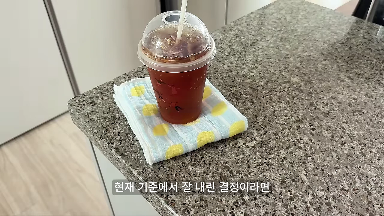 얼음이 담긴 플라스틱 일회용 커피 컵이 노란색과 하늘색 도트 무늬가 있는 작은 수건 위에 놓여 있고, 그 아래에는 '현재 기준에서 잘 내린 결정이라면'이라는 자막이 적혀 있습니다.