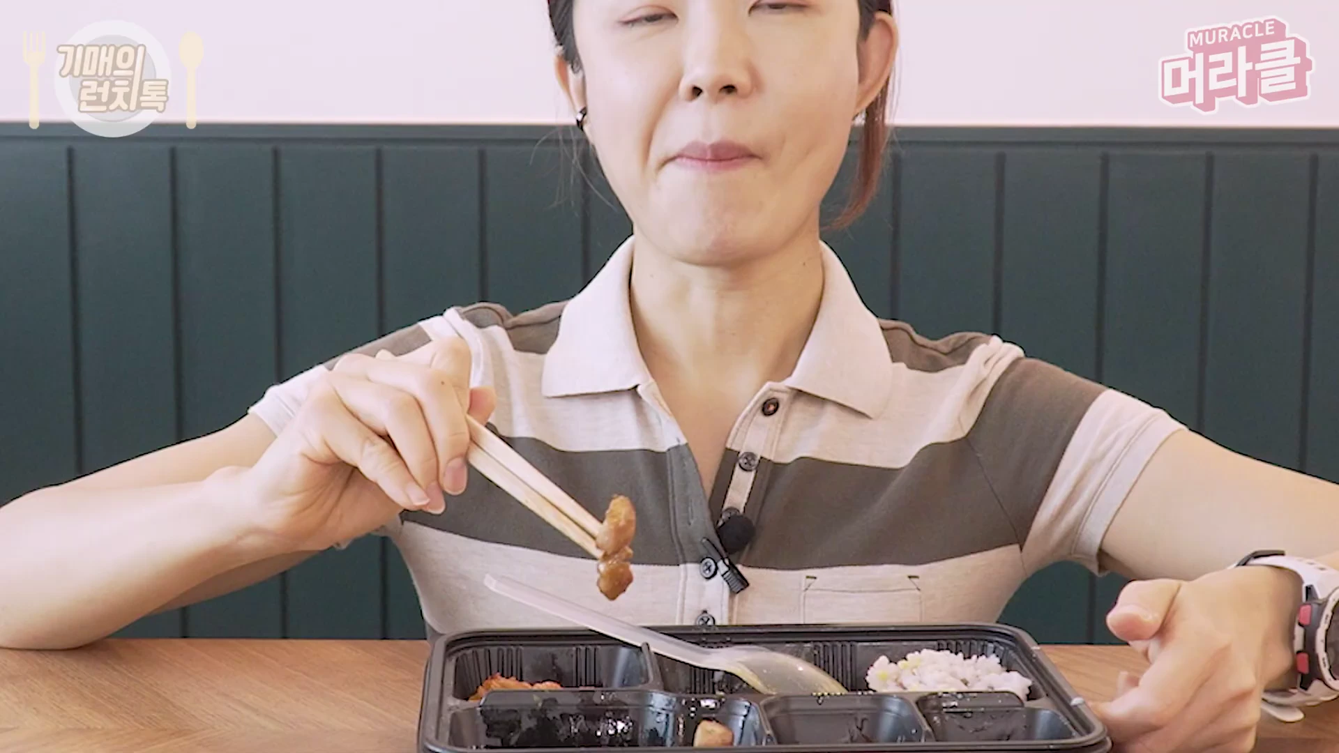줄무늬 셔츠를 입은 여성이 테이블 앞에서 젓가락으로 도시락 속 고기 반찬을 집어 먹고 있는 모습이다.
