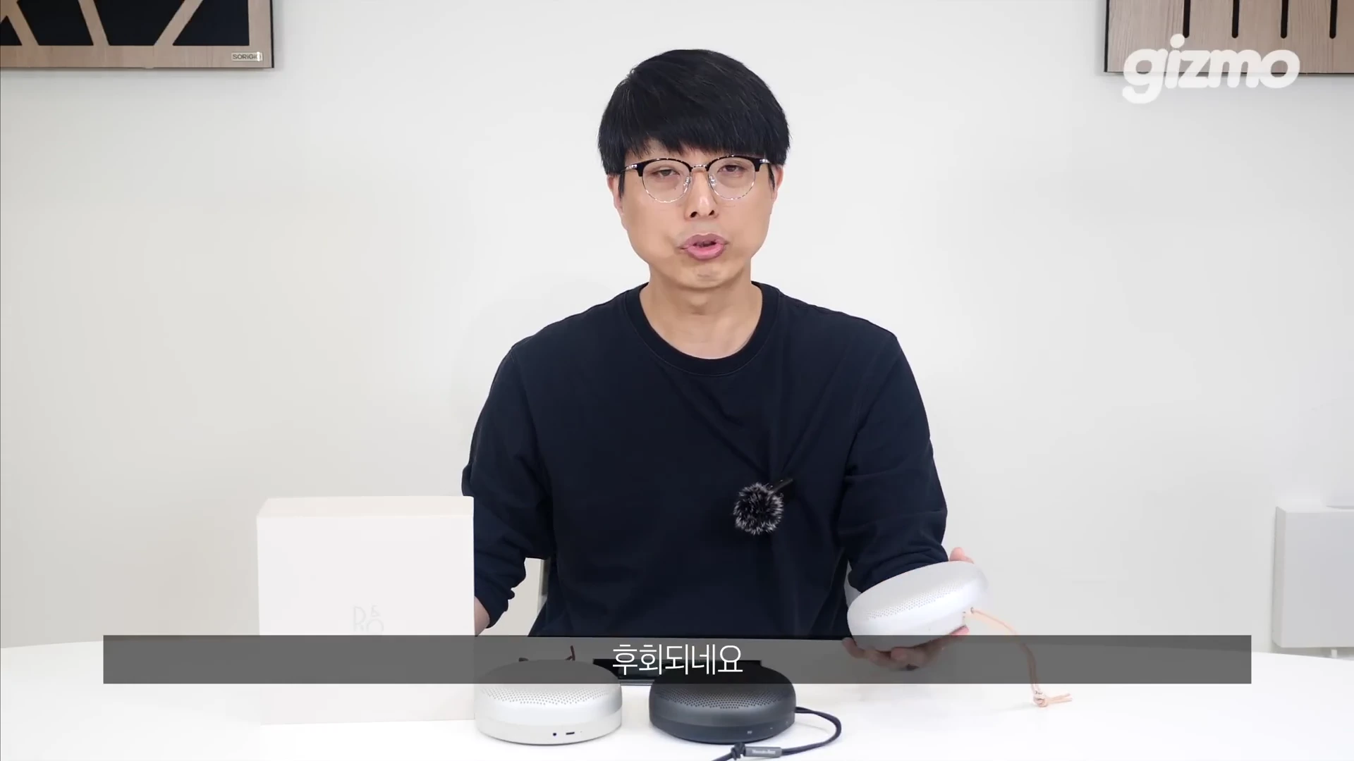 안경을 쓴 남성이 스튜디오 책상 앞에 앉아 뱅앤올룹슨 휴대용 스피커를 손에 들고 설명하고 있으며, 앞에는 스피커 박스와 두 대의 스피커가 놓여 있습니다.