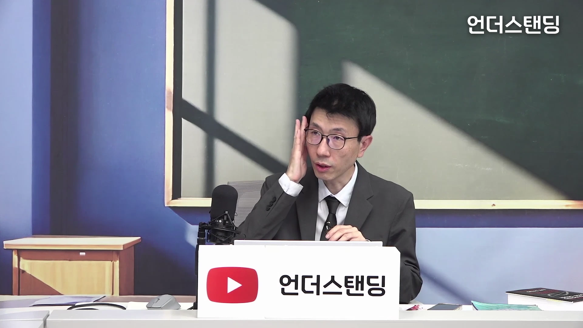 안경을 쓴 한 남성이 스튜디오 책상에 앉아 손으로 관자놀이 근처를 만지며 설명하고 있다.