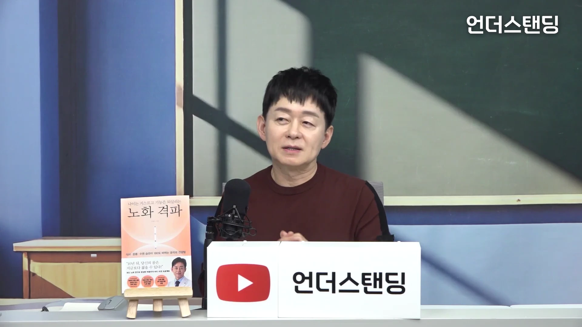 짙은 갈색 니트를 입은 남성이 스튜디오에서 마이크 앞에 앉아 이야기를 나누고 있으며, 옆에는 '노화 격파'라는 제목의 책이 놓여 있습니다.