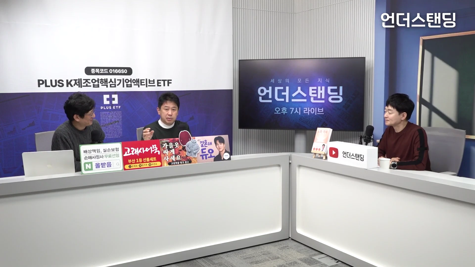 스튜디오에서 세 명의 남성이 책상에 앉아 대화를 나누는 모습으로, 앞에는 여러 광고 문구가 적힌 패널과 책이 놓여 있고 배경에는 대형 모니터가 설치되어 있습니다.