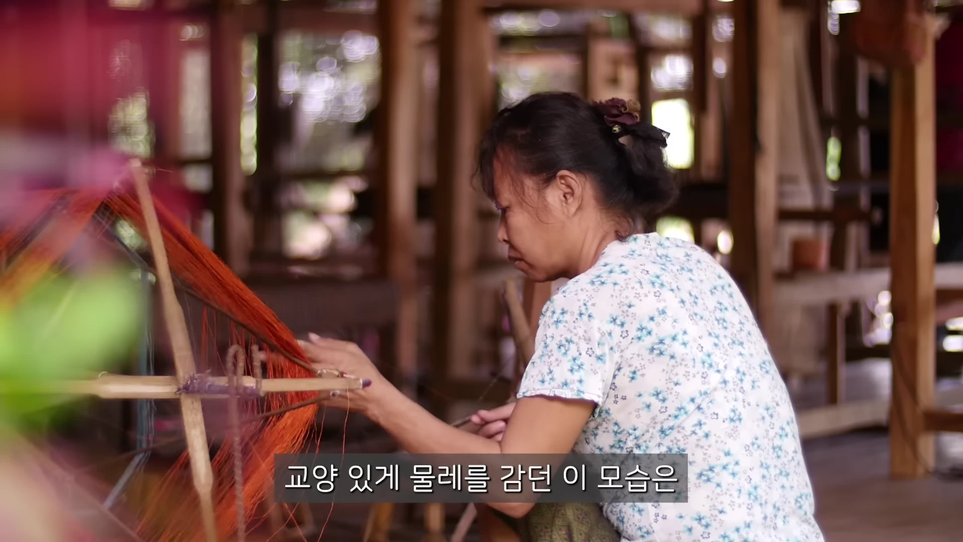 전통 복장을 한 여성이 나무 틀에 감긴 주황색 실을 손으로 정리하고 있는 모습이다.