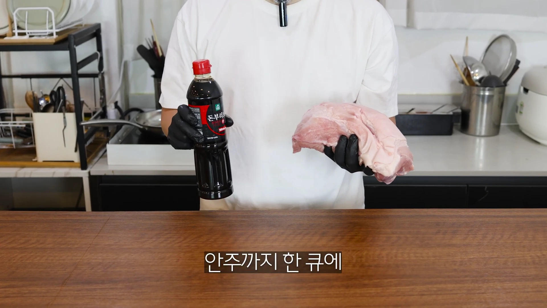 흰 티셔츠를 입고 검은색 위생 장갑을 낀 사람이 한 손에는 소스 병을, 다른 한 손에는 생고기 덩어리를 들고 서 있다.