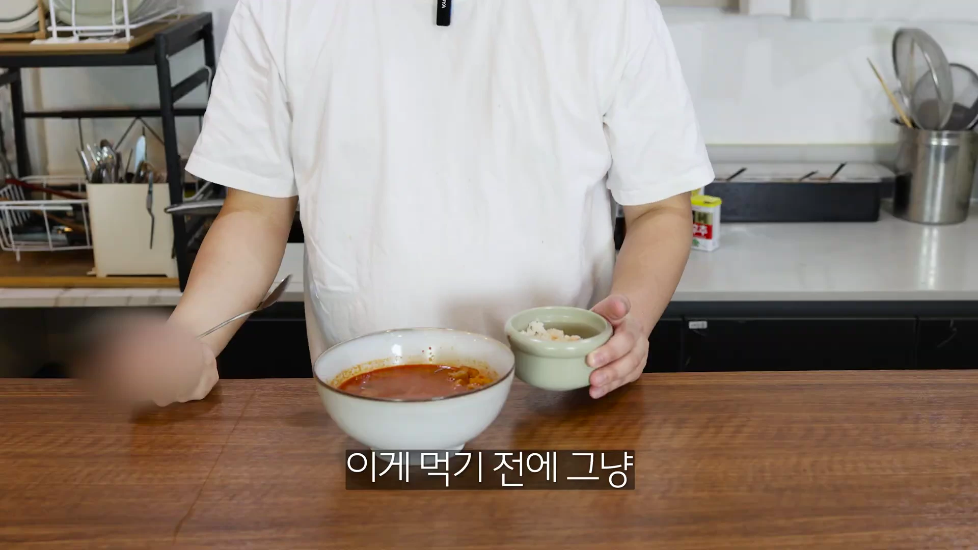 흰 티셔츠를 입은 사람이 한 손에는 붉은 국물 요리가 담긴 그릇을, 다른 한 손에는 밥이 담긴 작은 그릇을 들고 있는 모습.