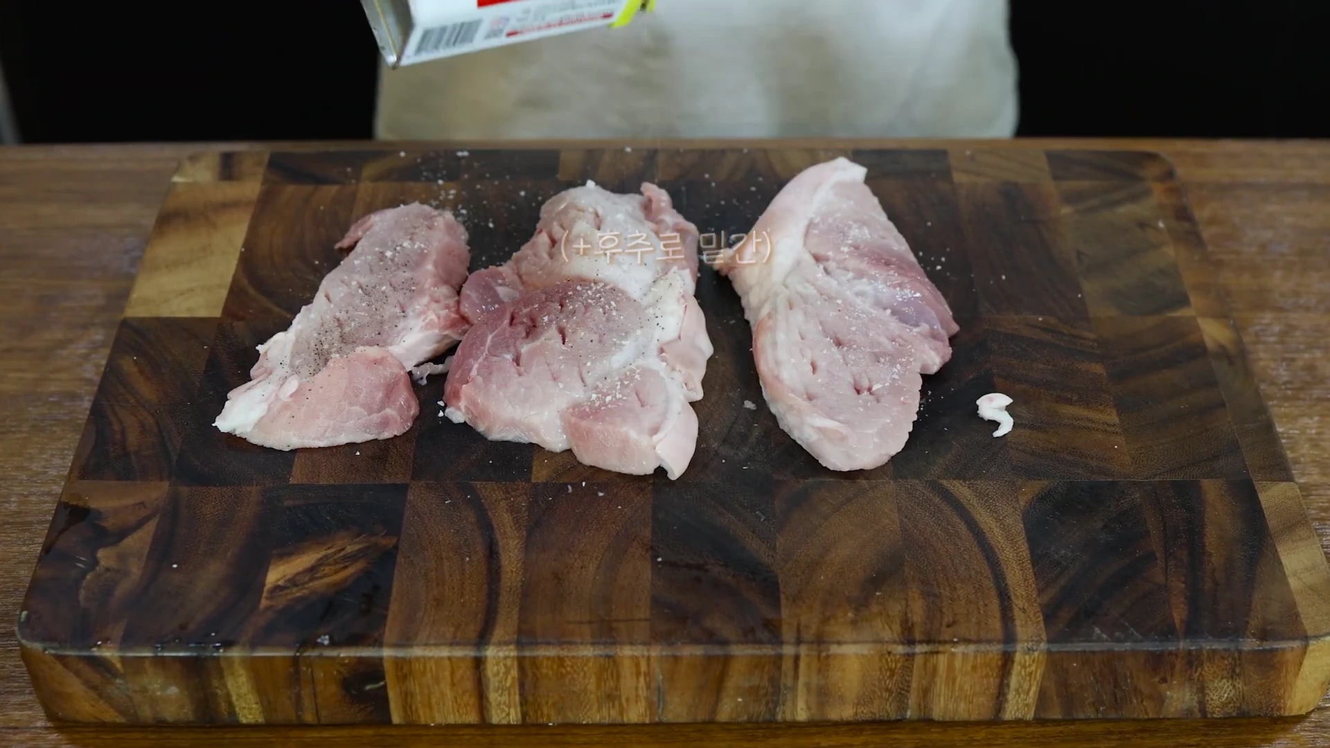 나무 도마 위에 올려진 돼지고기 뒷다리살 덩어리들에 소금과 후추를 뿌리고 있는 모습.
