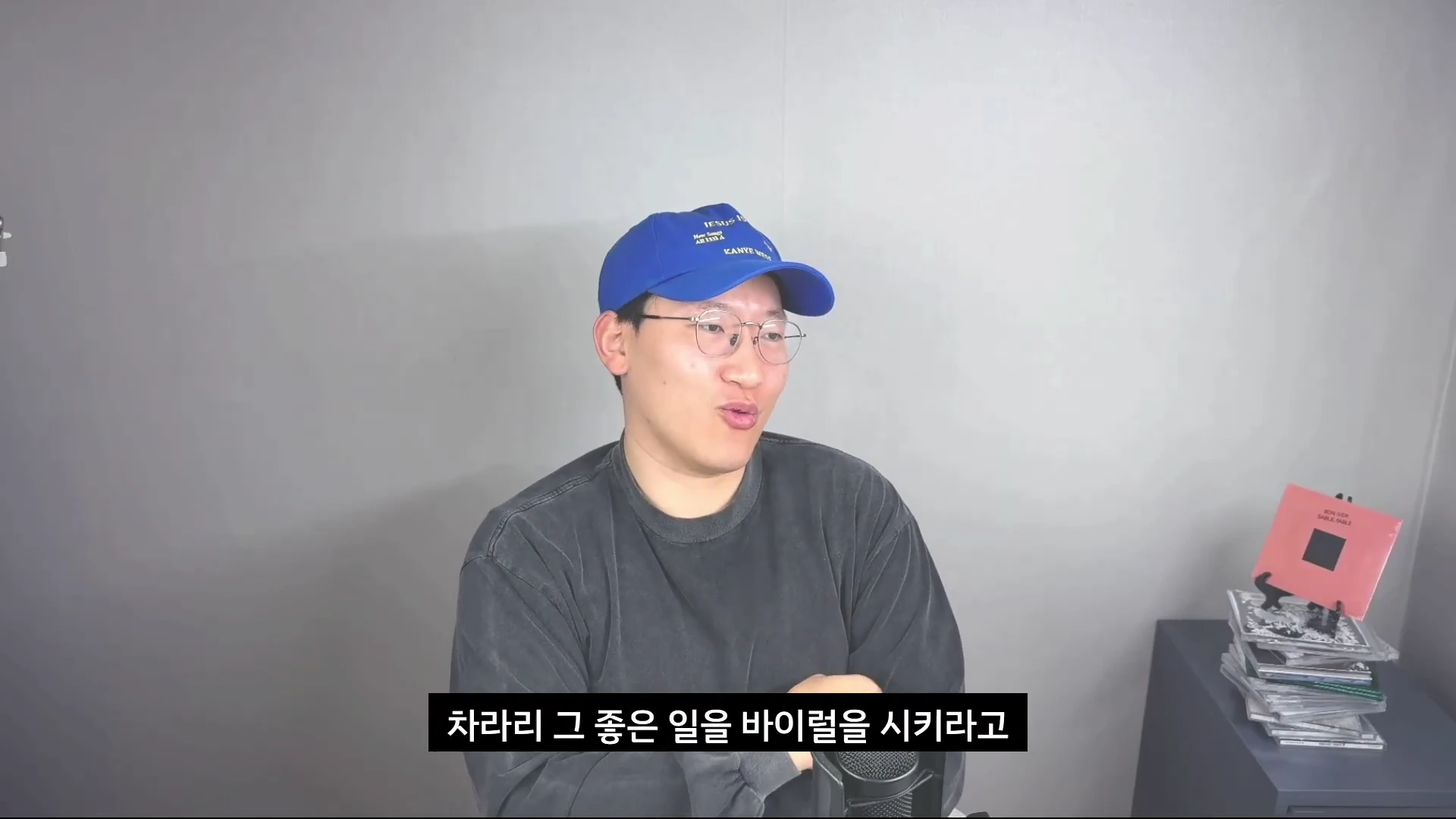 파란색 야구 모자와 안경을 쓴 남성이 마이크 앞에 앉아 정면을 바라보며 이야기하고 있는 모습입니다.