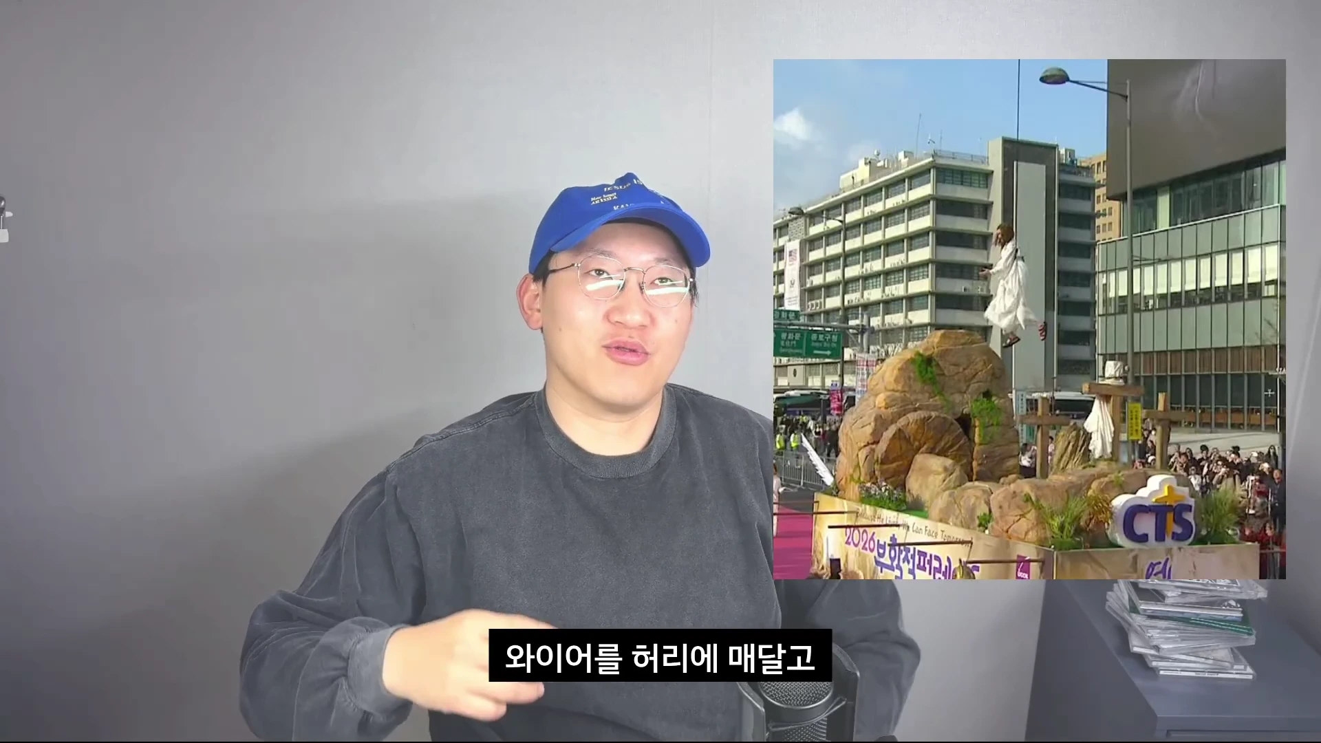 파란 모자를 쓴 남성이 스튜디오에서 이야기하고 있고, 화면 오른쪽에는 거리 퍼레이드에서 흰 옷을 입은 배우가 와이어에 매달려 공중으로 올라가는 모습이 담긴 영상이 삽입되어 있습니다.