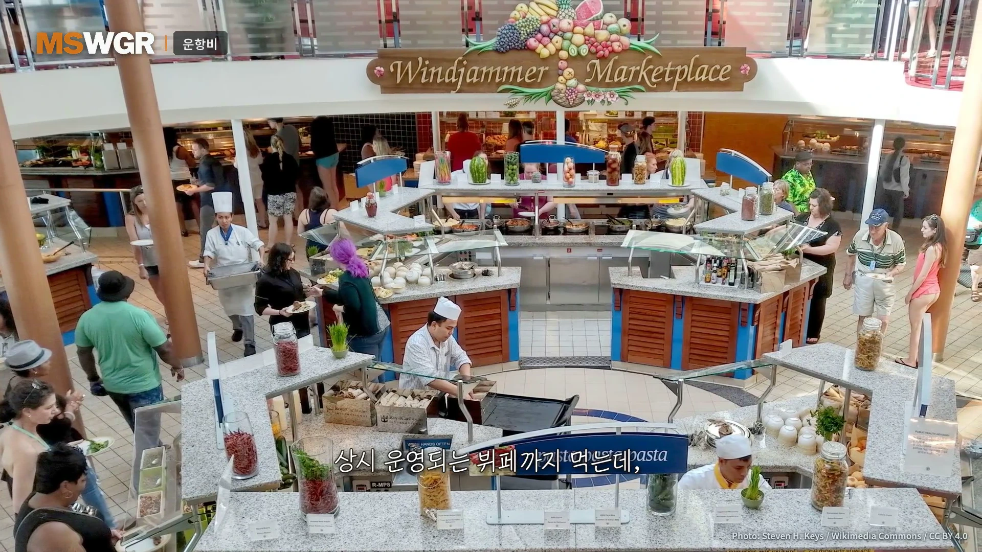 크루즈 선박 내의 뷔페 식당 모습으로, 승객들이 여러 조리대 사이를 오가며 음식을 담고 있고 요리사들이 서비스를 제공하고 있습니다.