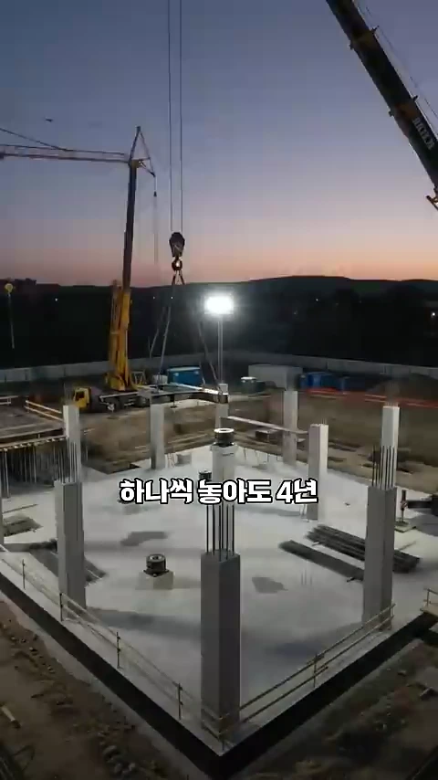 공사 현장의 지하 기초 위에 여러 개의 기둥이 세워져 있고 크레인이 자재를 들어 올리는 모습이 촬영된 조감도입니다.