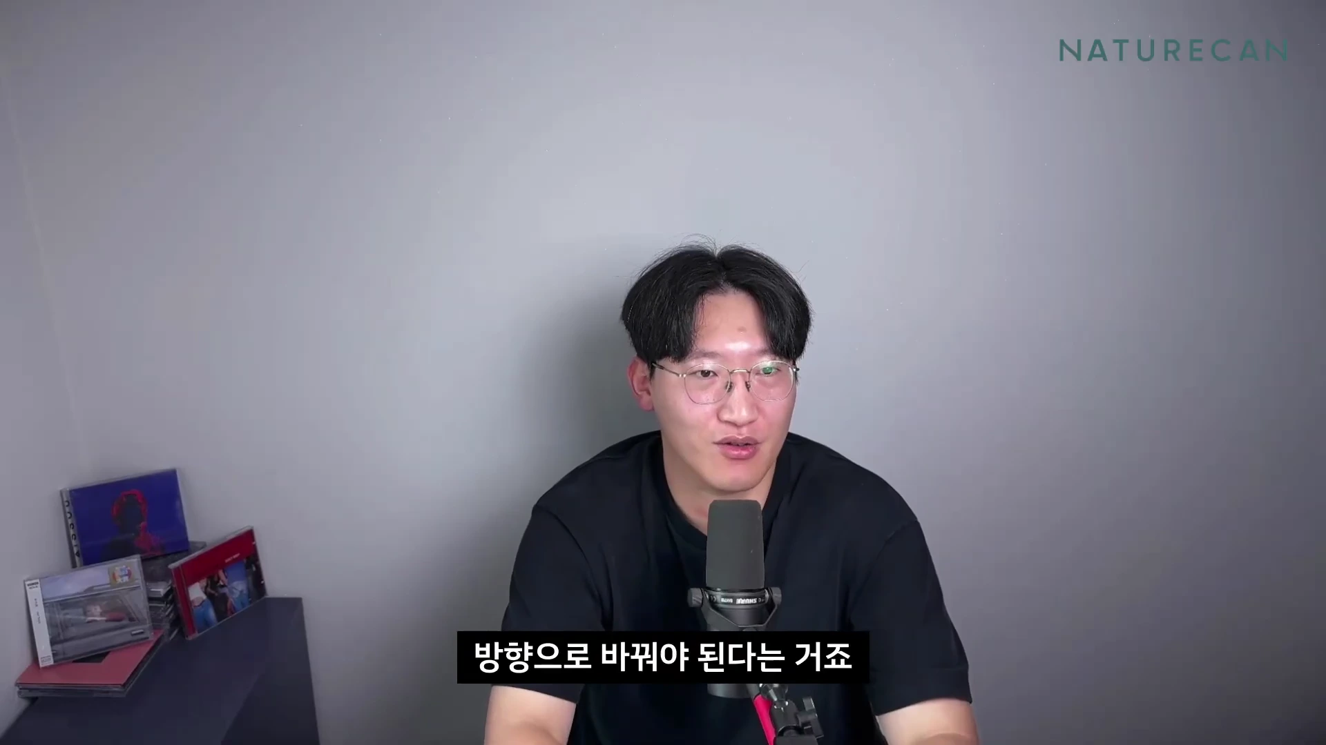 검은색 반팔 티셔츠를 입고 안경을 쓴 남성이 마이크 앞에서 이야기하고 있으며 화면 하단에는 '방향으로 바꿔야 된다는 거죠'라는 자막이 표시되어 있습니다.