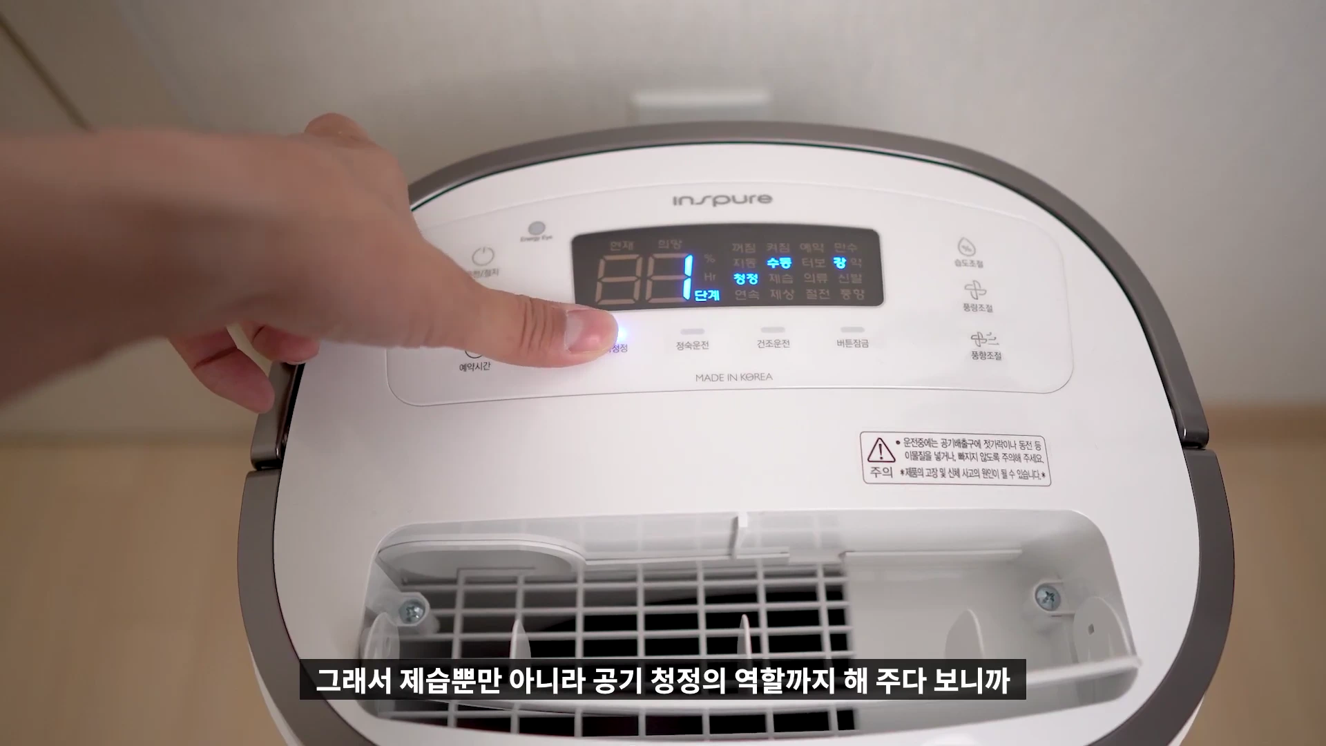 사람의 손가락이 하얀색 공기청정 제습기의 상단 제어판 버튼을 누르고 있으며, LED 화면에는 현재 상태와 설정 메뉴가 표시되어 있습니다.