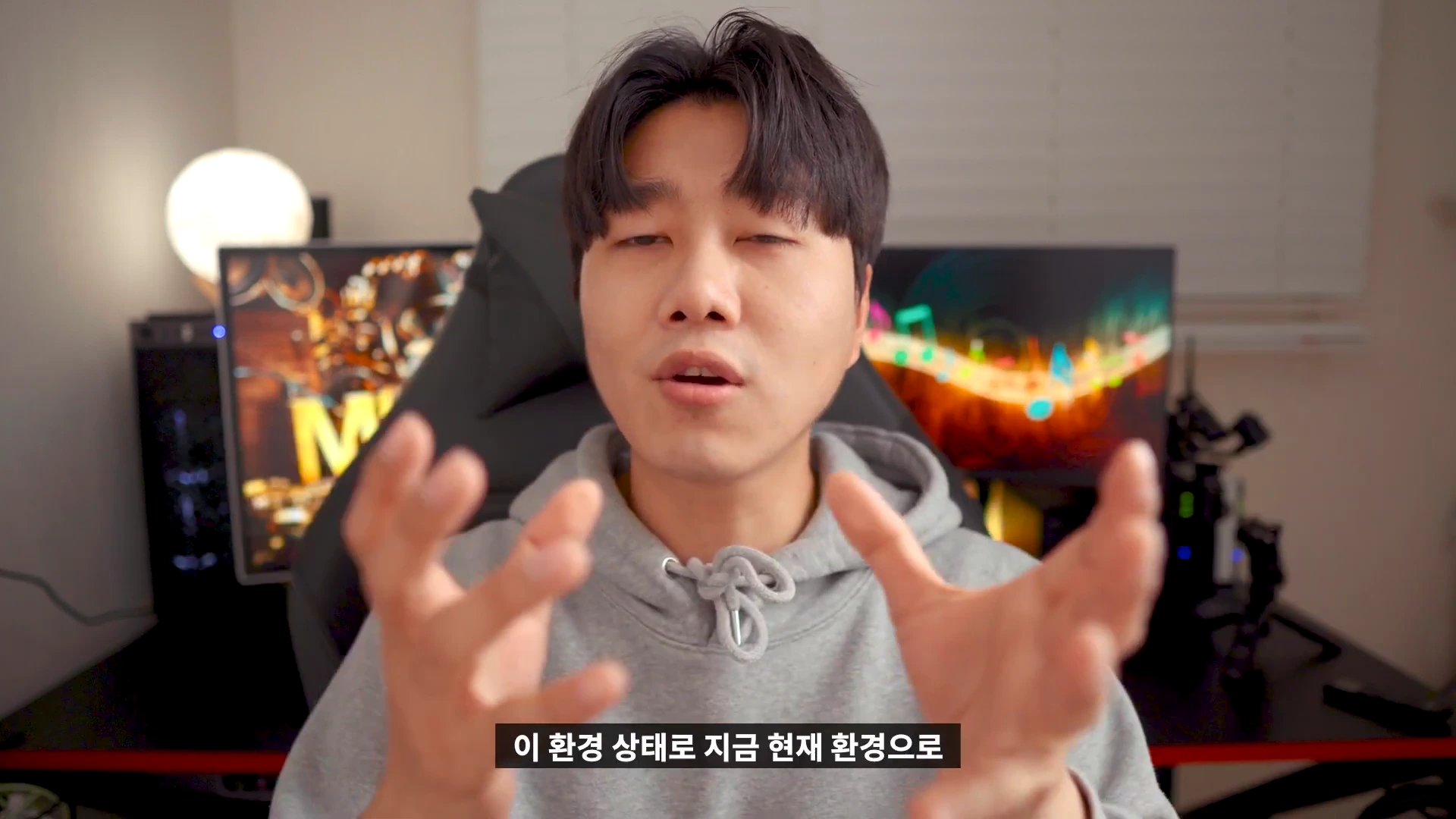 회색 후드티를 입은 남성이 정면을 바라보며 손을 흔들며 이야기를 하고 있는 모습