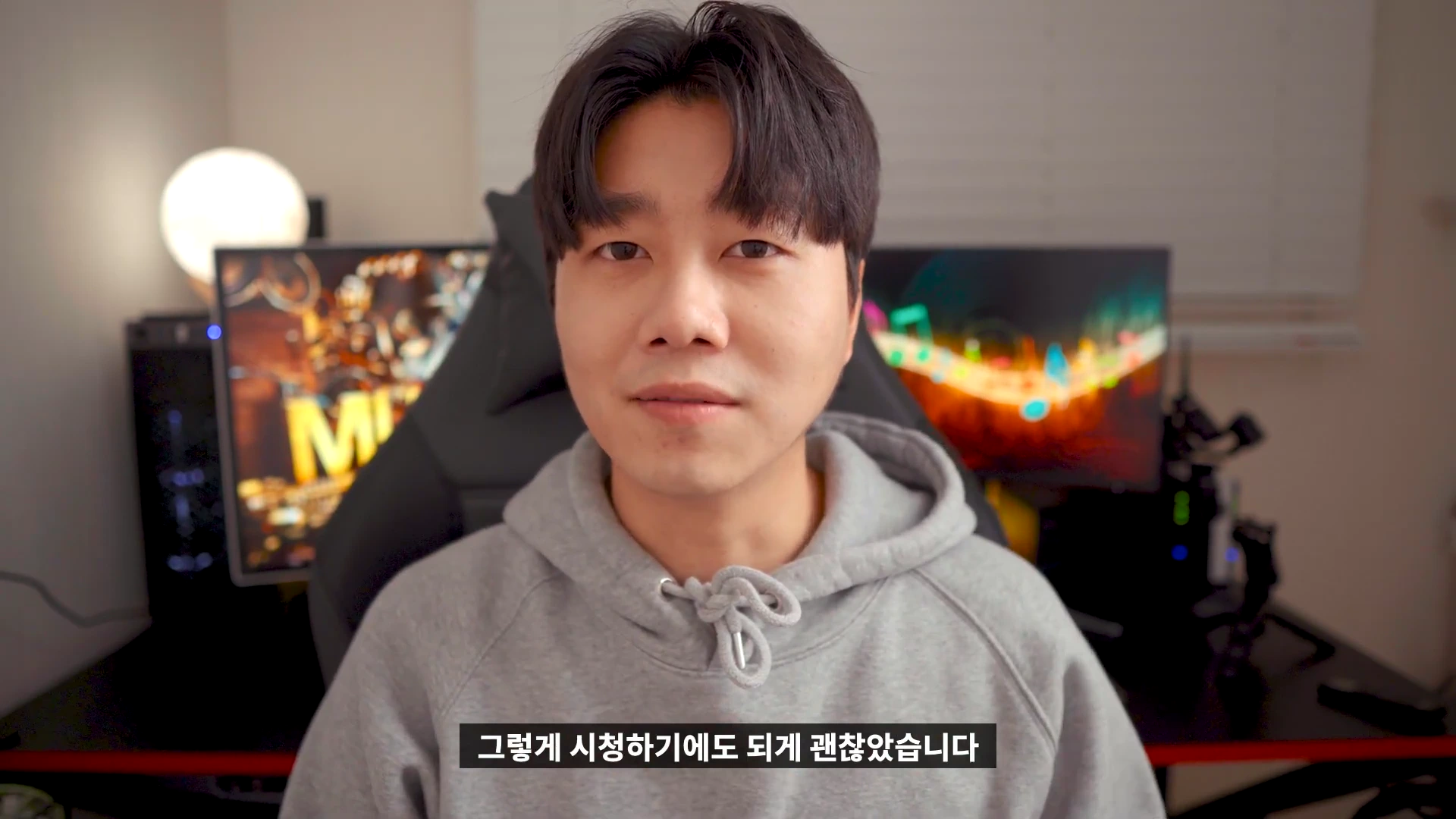 실내에서 회색 후드티를 입은 남성이 정면을 응시하며 이야기하고 있는 모습