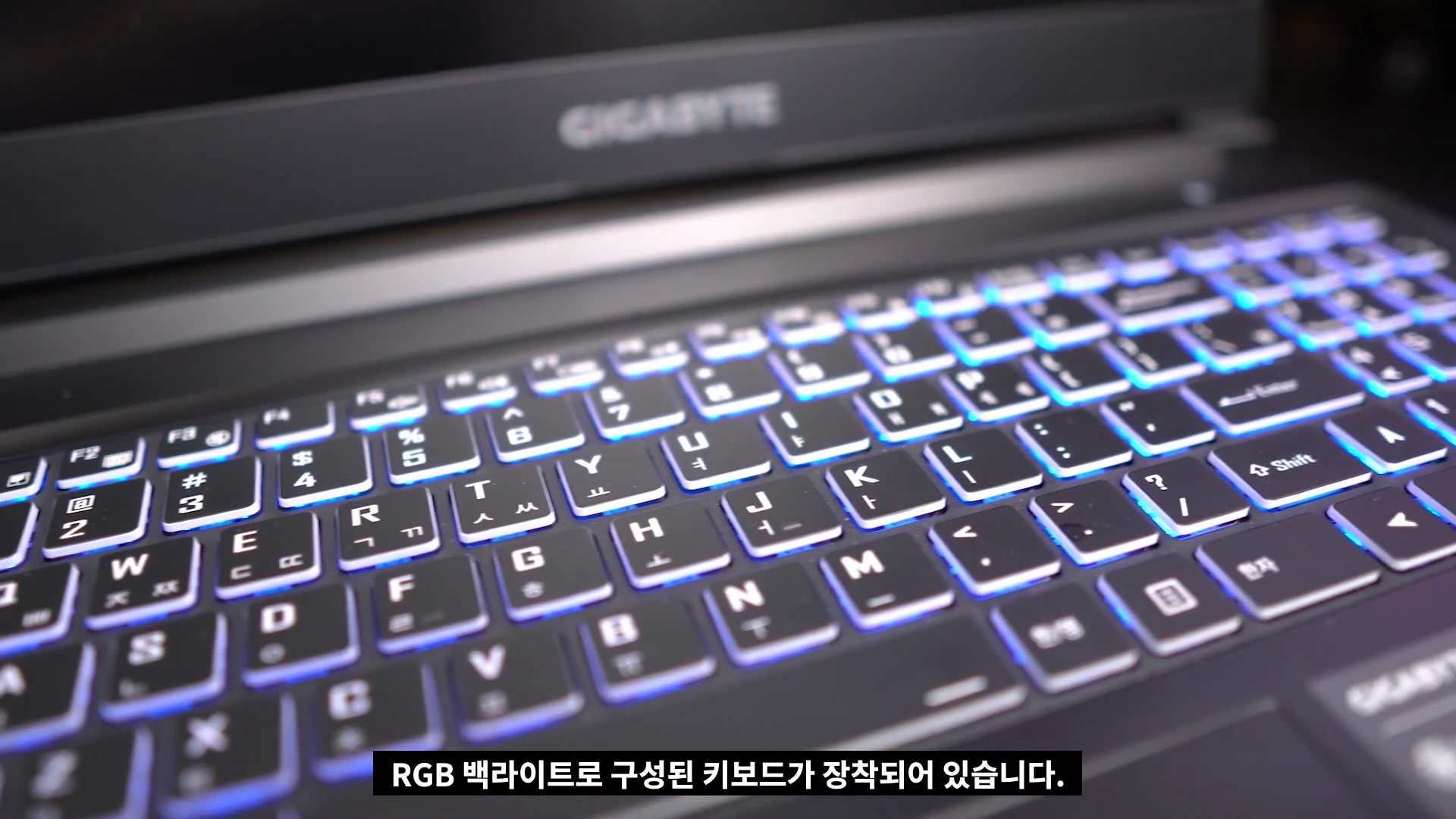 노트북 키보드 상단부와 GIGABYTE 로고가 보이는 클로즈업 사진으로, 키보드 아래에서 파란색 RGB 조명이 밝게 빛나고 있습니다.