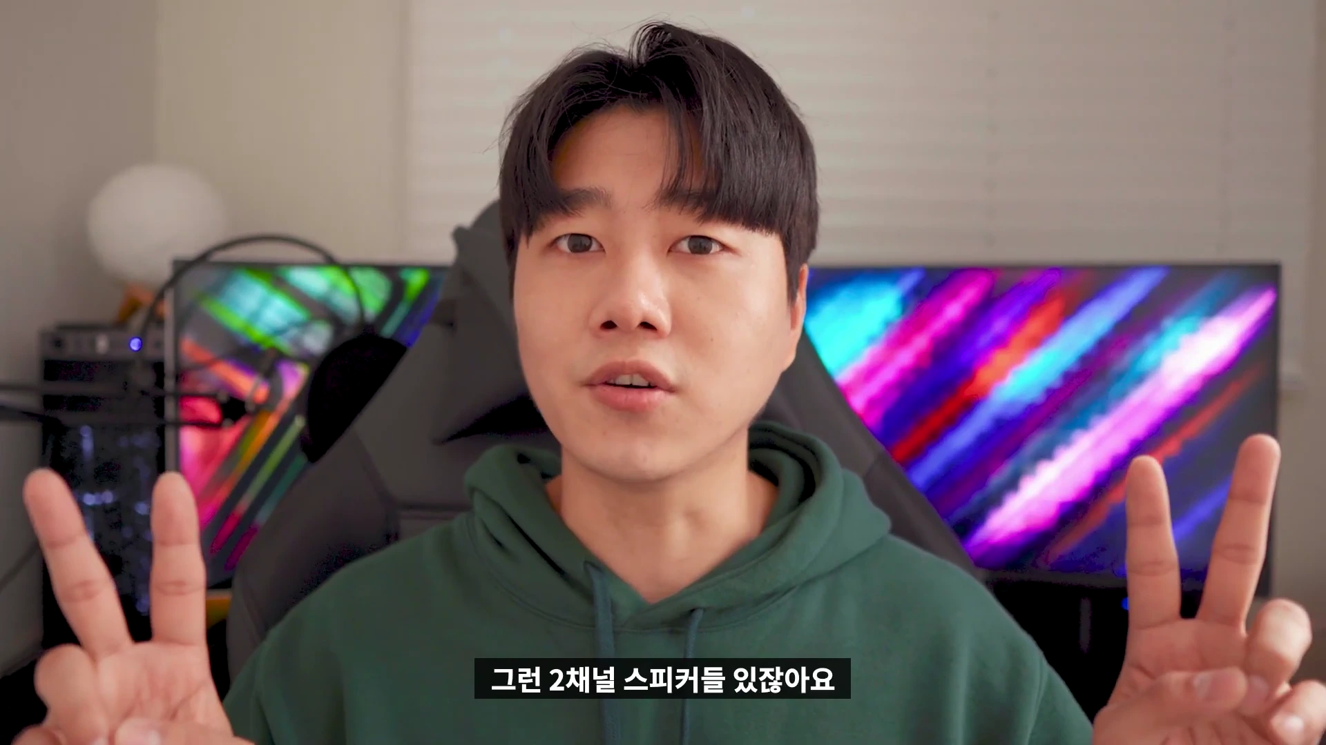 녹색 후드티를 입은 남성이 카메라를 보며 양손으로 V자를 그리고 있는 상반신 샷