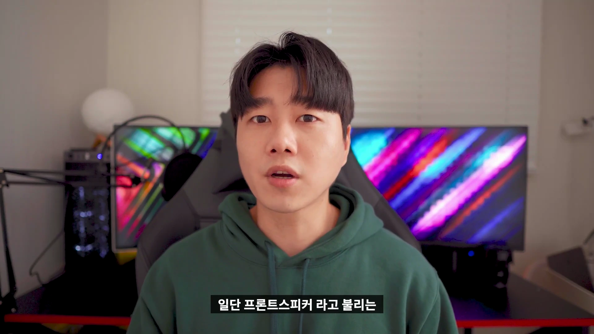 초록색 후드티를 입은 남성이 카메라를 보며 이야기를 하고 있는 상반신 샷으로, 배경에는 화려한 색감의 모니터 화면이 보입니다.