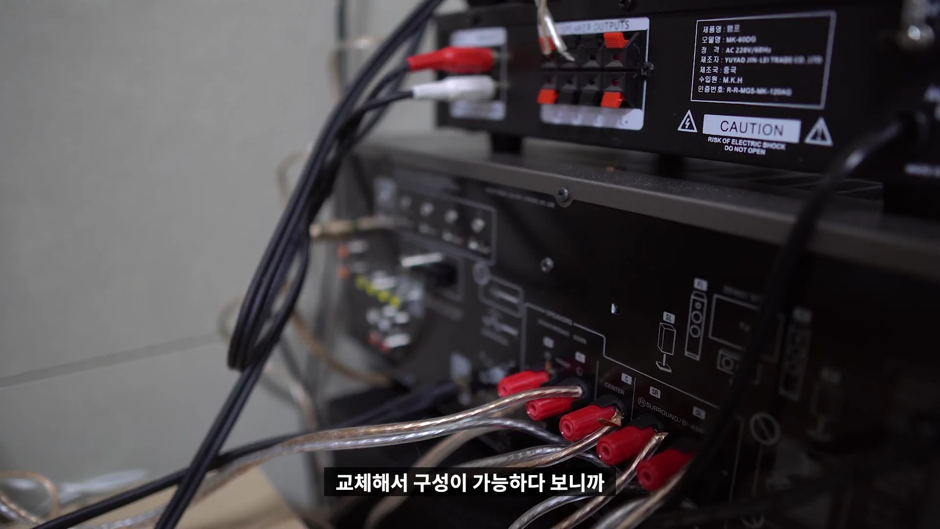 오디오 앰프 뒷면의 여러 케이블 연결 단자와 스피커 출력 단자가 보이고, 빨간색과 검은색 커넥터에 투명한 스피커 선들이 꽂혀 있는 모습입니다.