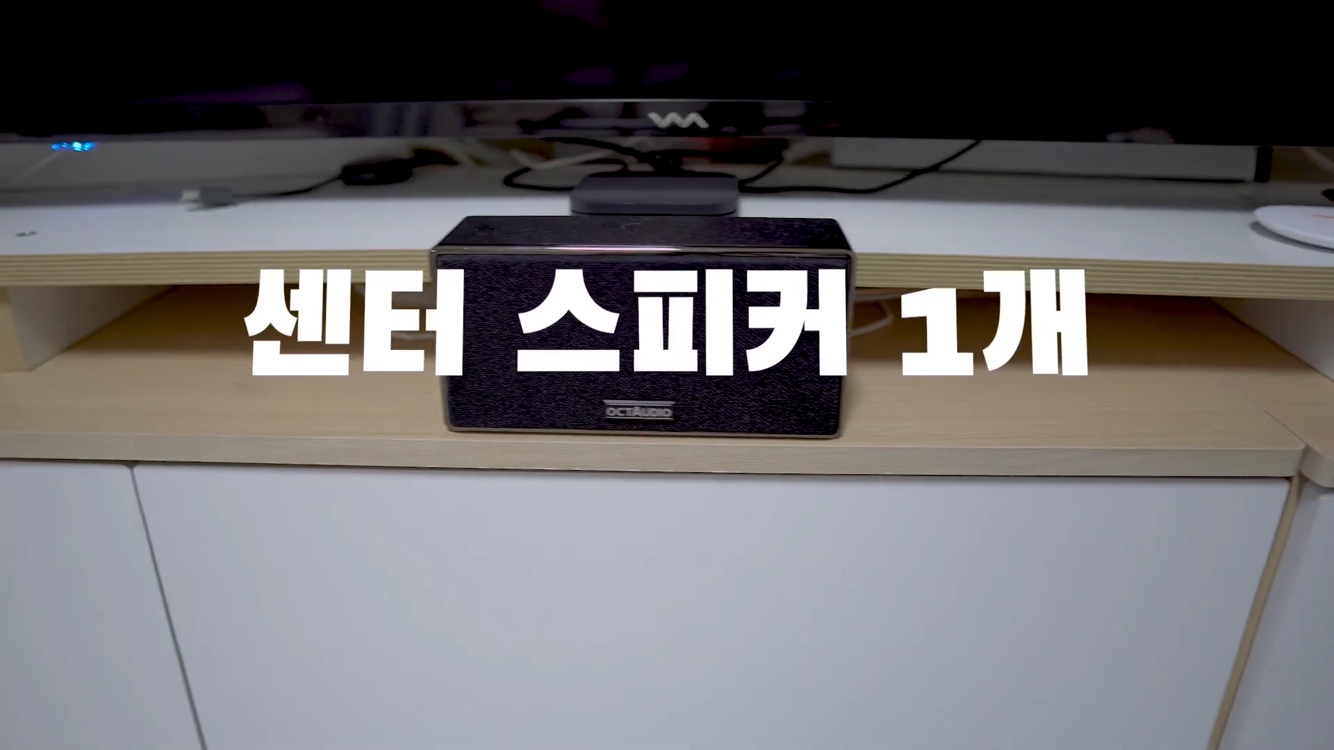 흰색 TV 장식장 위에 놓인 검은색 센터 스피커가 보이며 화면 중앙에는 흰색 자막으로 센터 스피커 1개라고 적혀 있습니다.