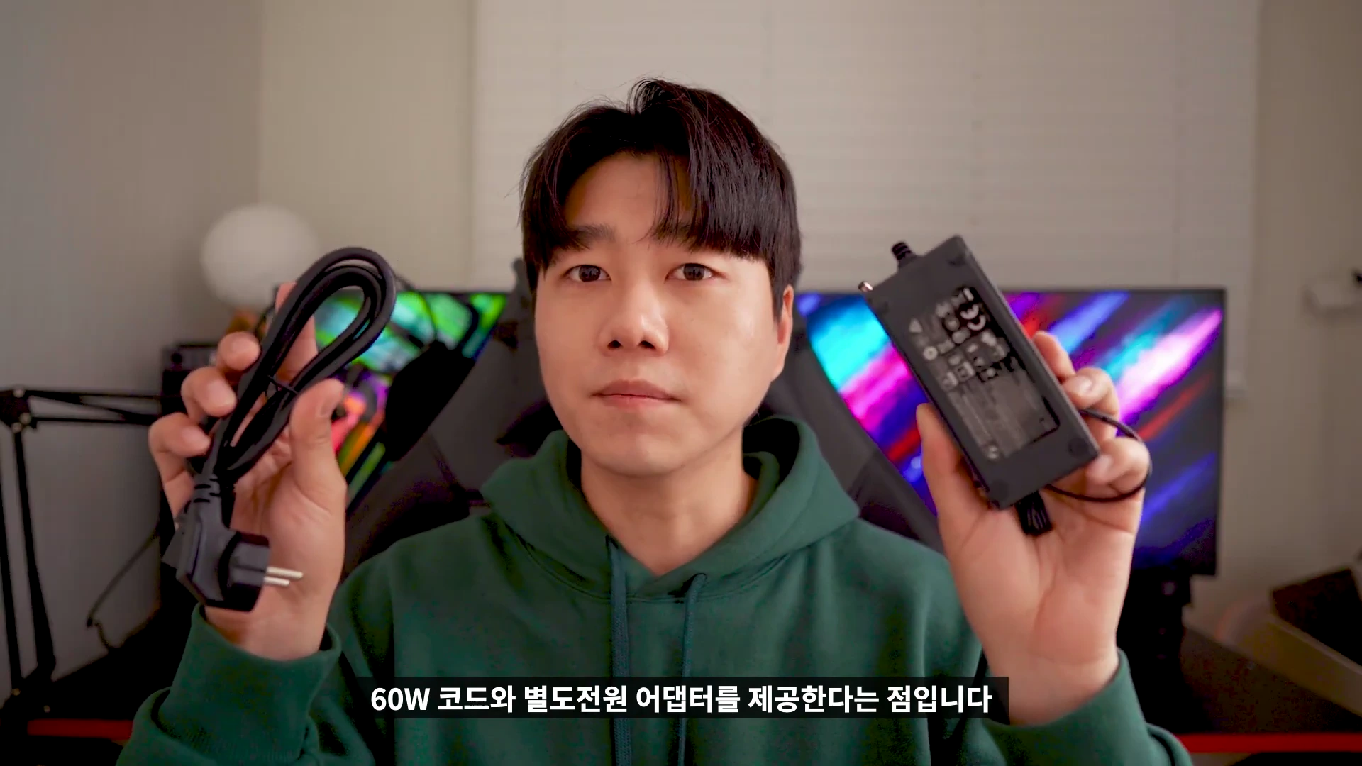 실내에서 녹색 후드티를 입은 남성이 양손에 60W 전원 어댑터와 전원 케이블을 들고 카메라를 응시하고 있습니다.