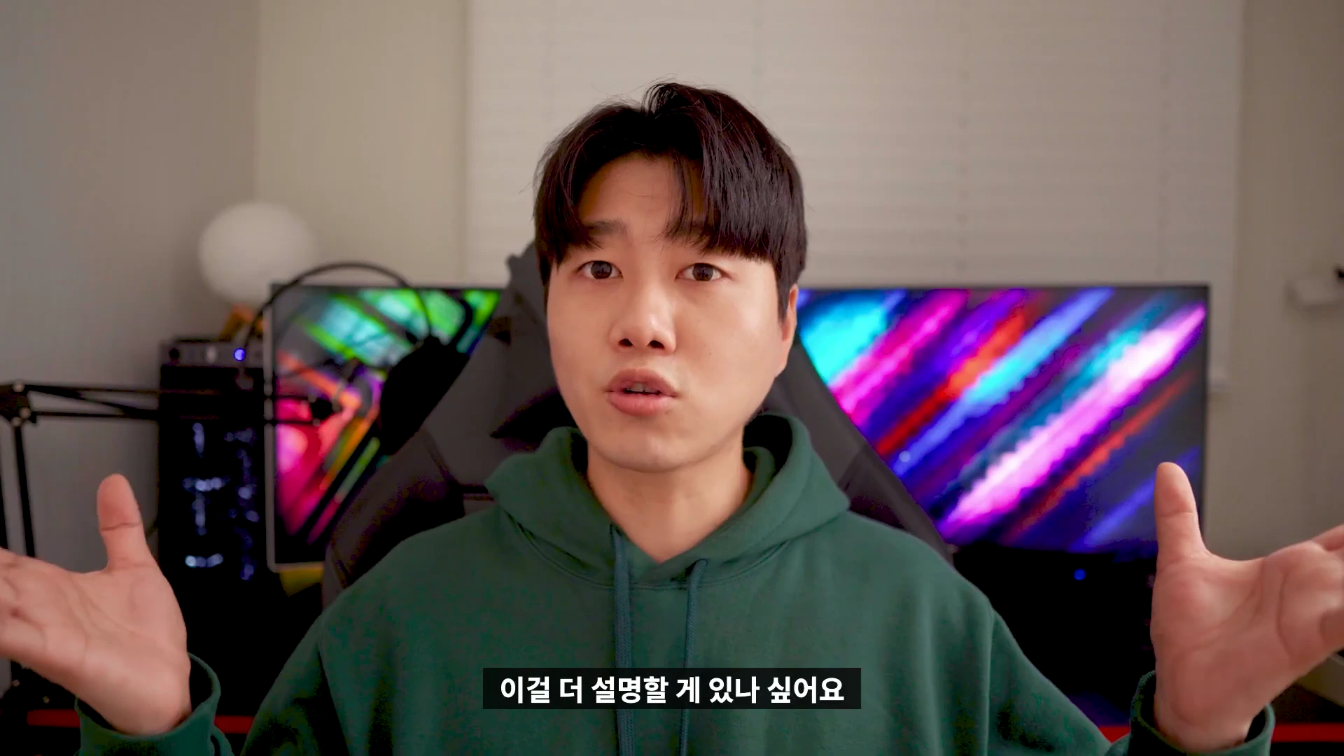 실내에서 초록색 후드티를 입은 남성이 양손을 벌리며 카메라를 정면으로 바라보고 있습니다.