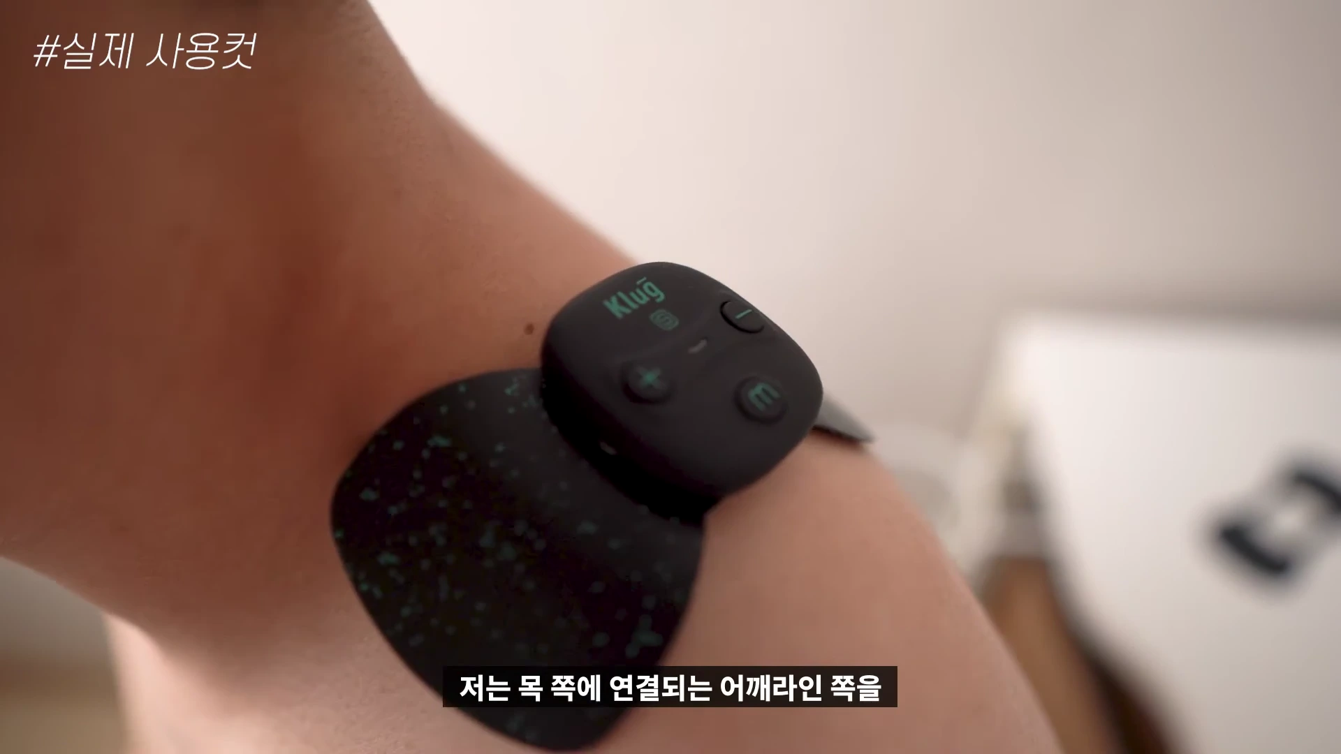 사람의 어깨 부위에 검은색의 작은 무선 저주파 마사지기가 부착되어 있는 근접 촬영 영상 화면입니다.